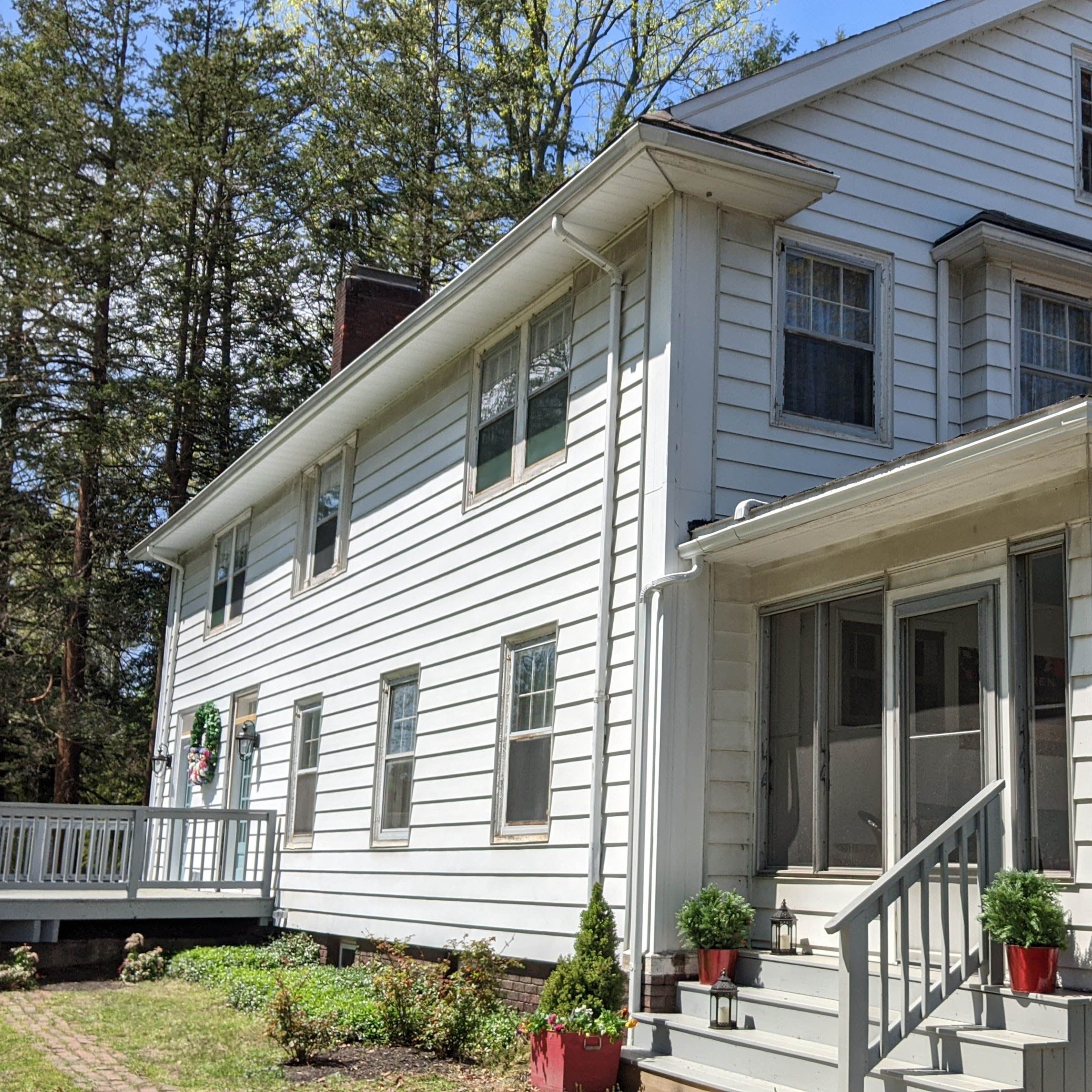 A two-story, white wooden house features a front porch and a side deck, surrounded by greenery.