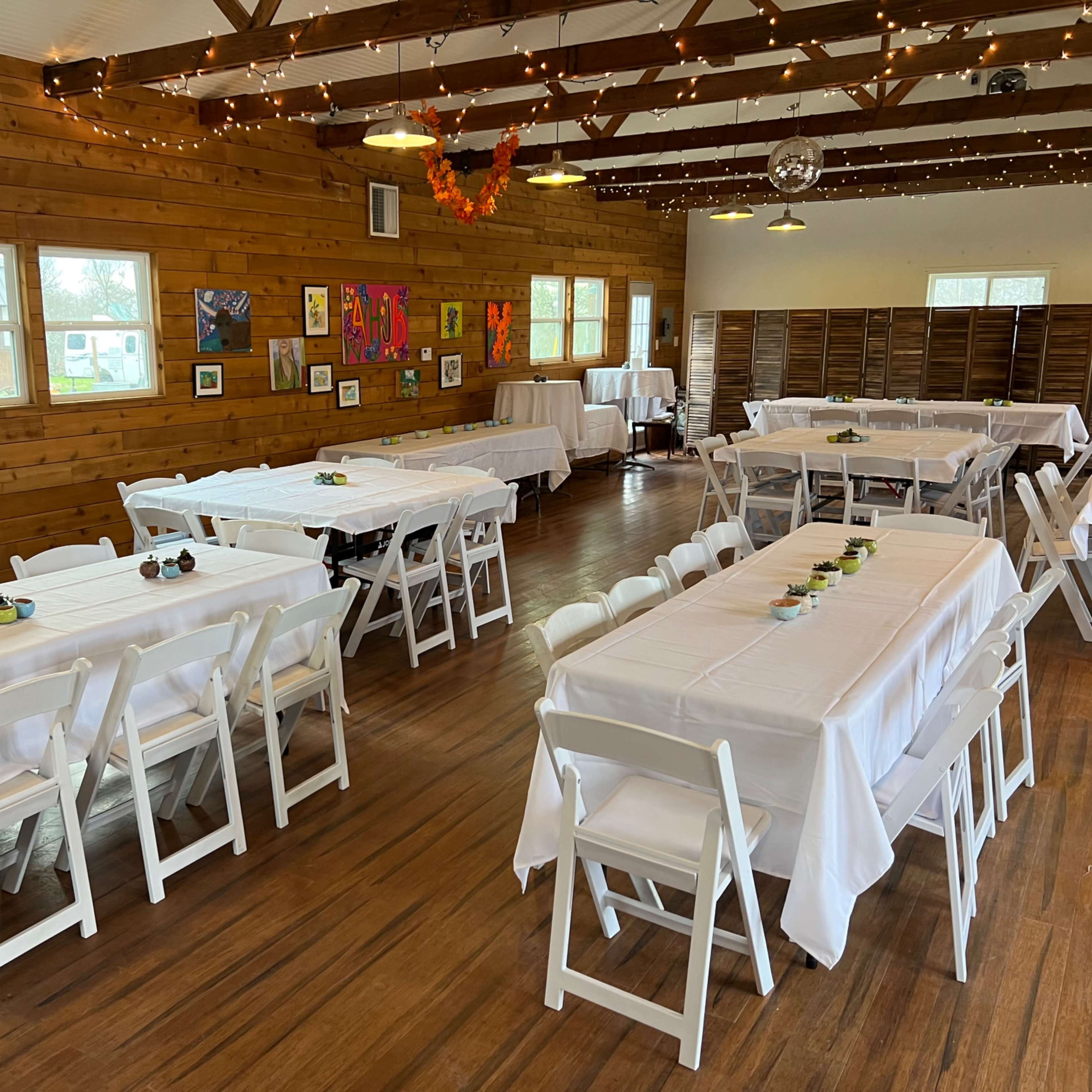 The interior of a banquet hall features neatly arranged tables with white tablecloths and wooden walls adorned with artwork.