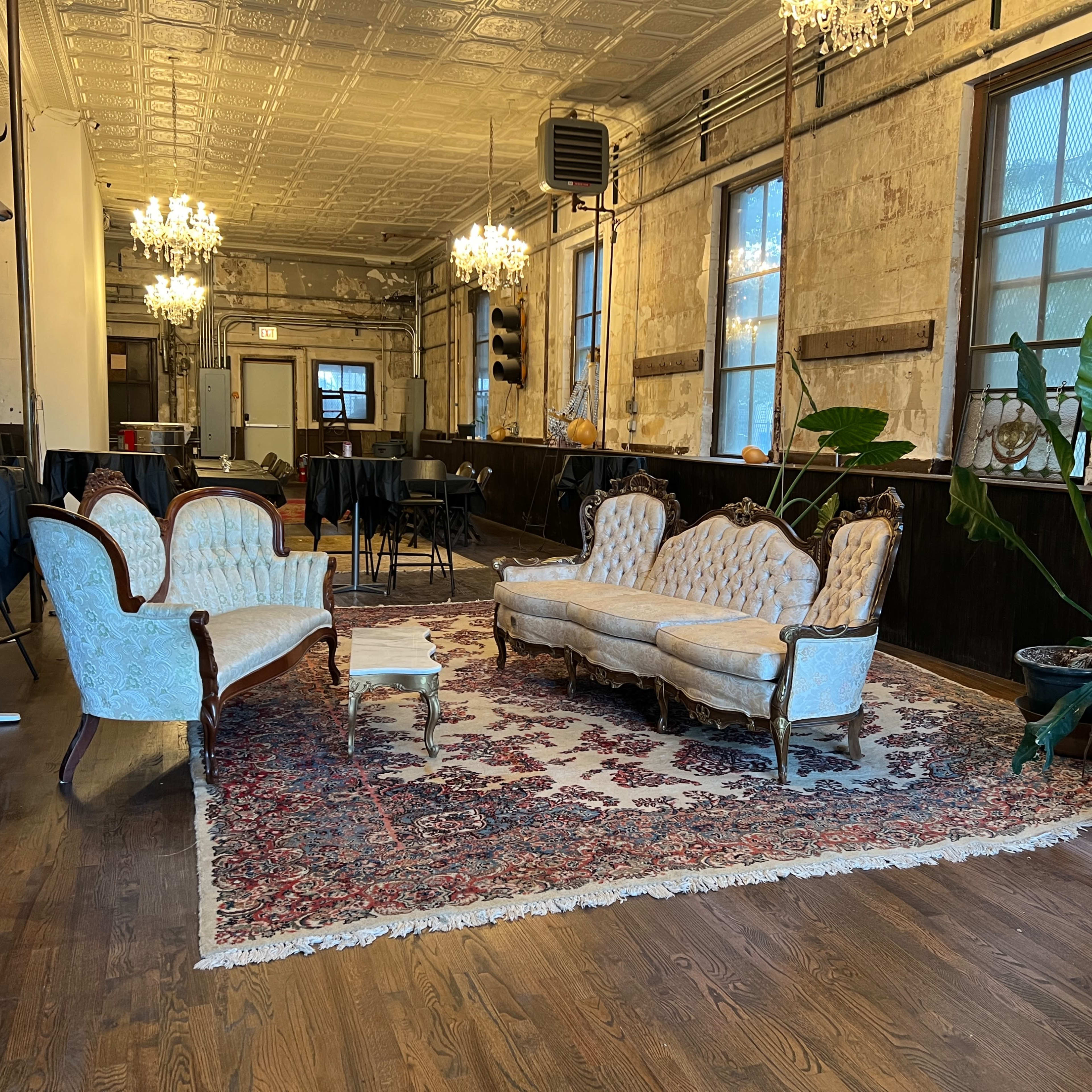 A vintage-style seating area with ornate furniture, a large patterned rug, and potted plants in a spacious room with exposed brick walls and chandeliers.