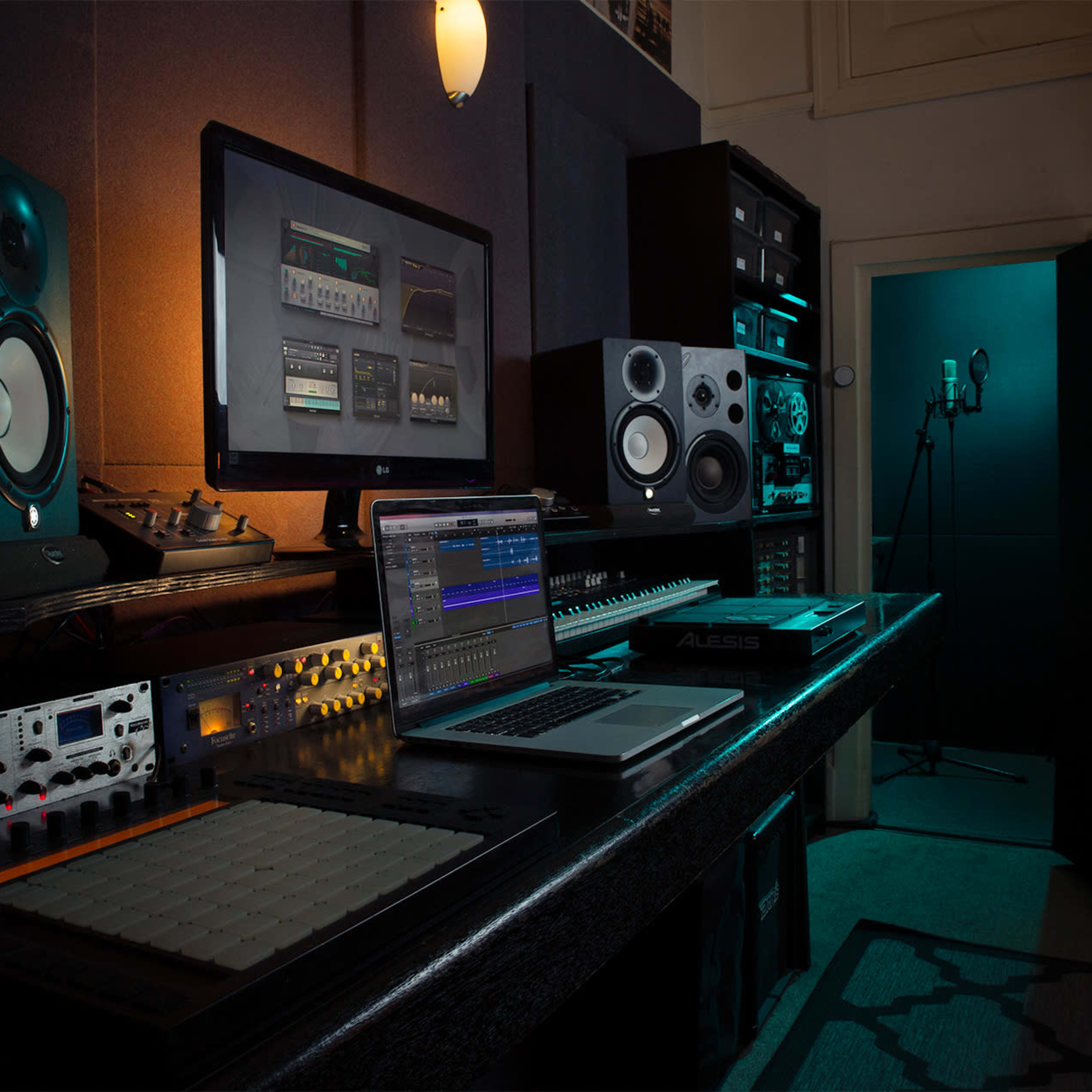 A music production studio contains a computer, audio interface, speakers, and a microphone set up in a dimly lit space.