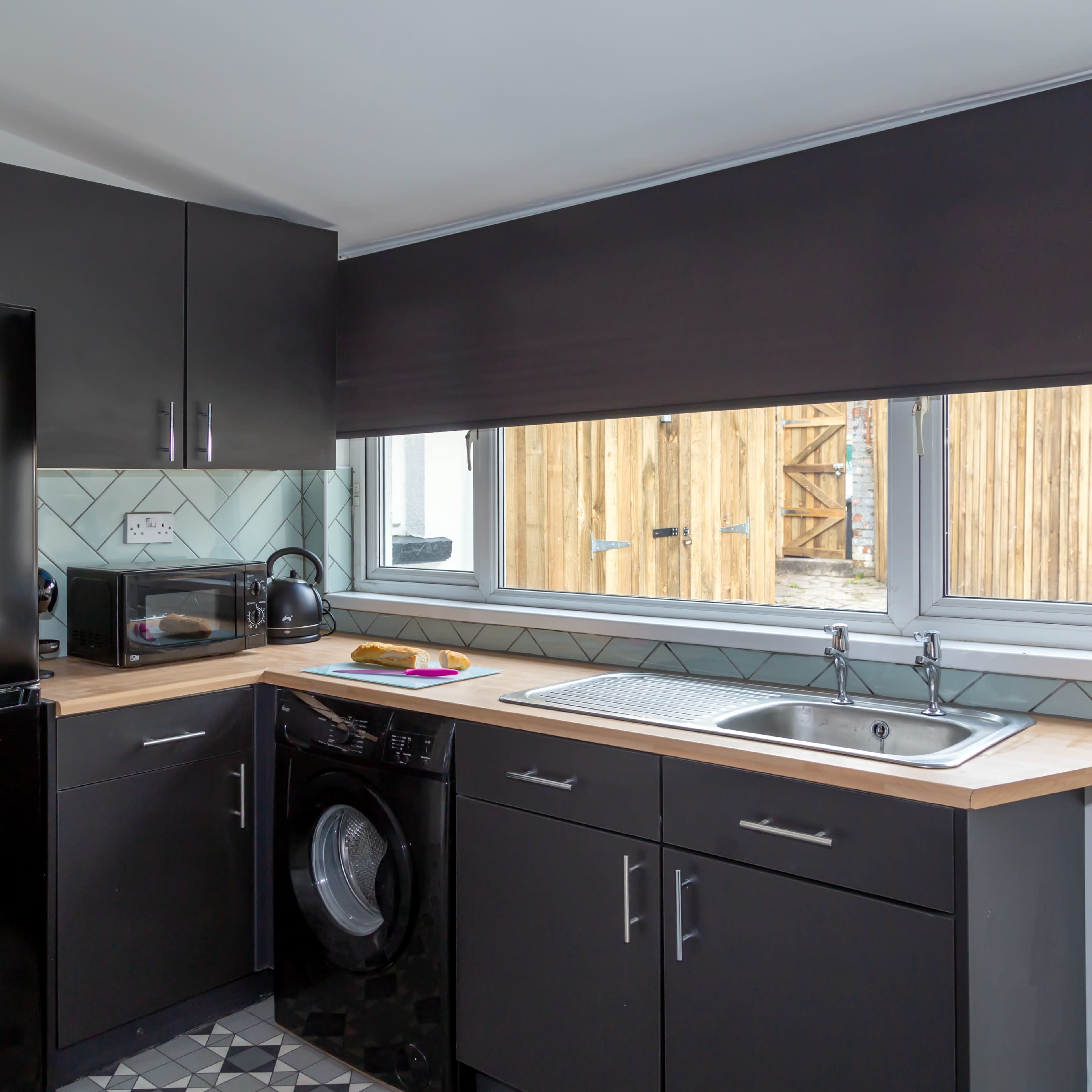 A modern kitchen with black cabinets, a black refrigerator, a washing machine, and a countertop under a large window.