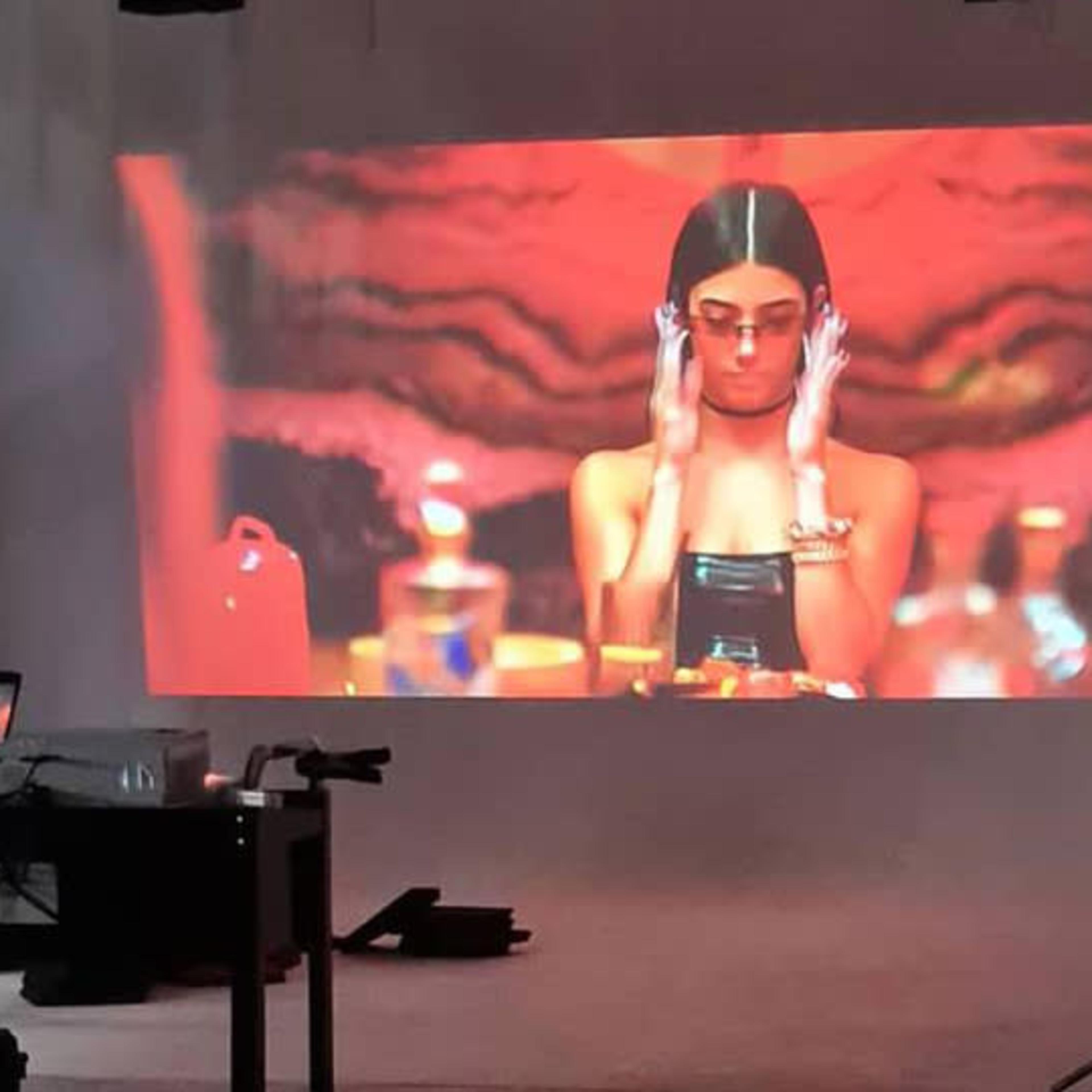 A large projection of a woman wearing sunglasses is displayed on a wall in a dimly lit room with various bottles on a table.