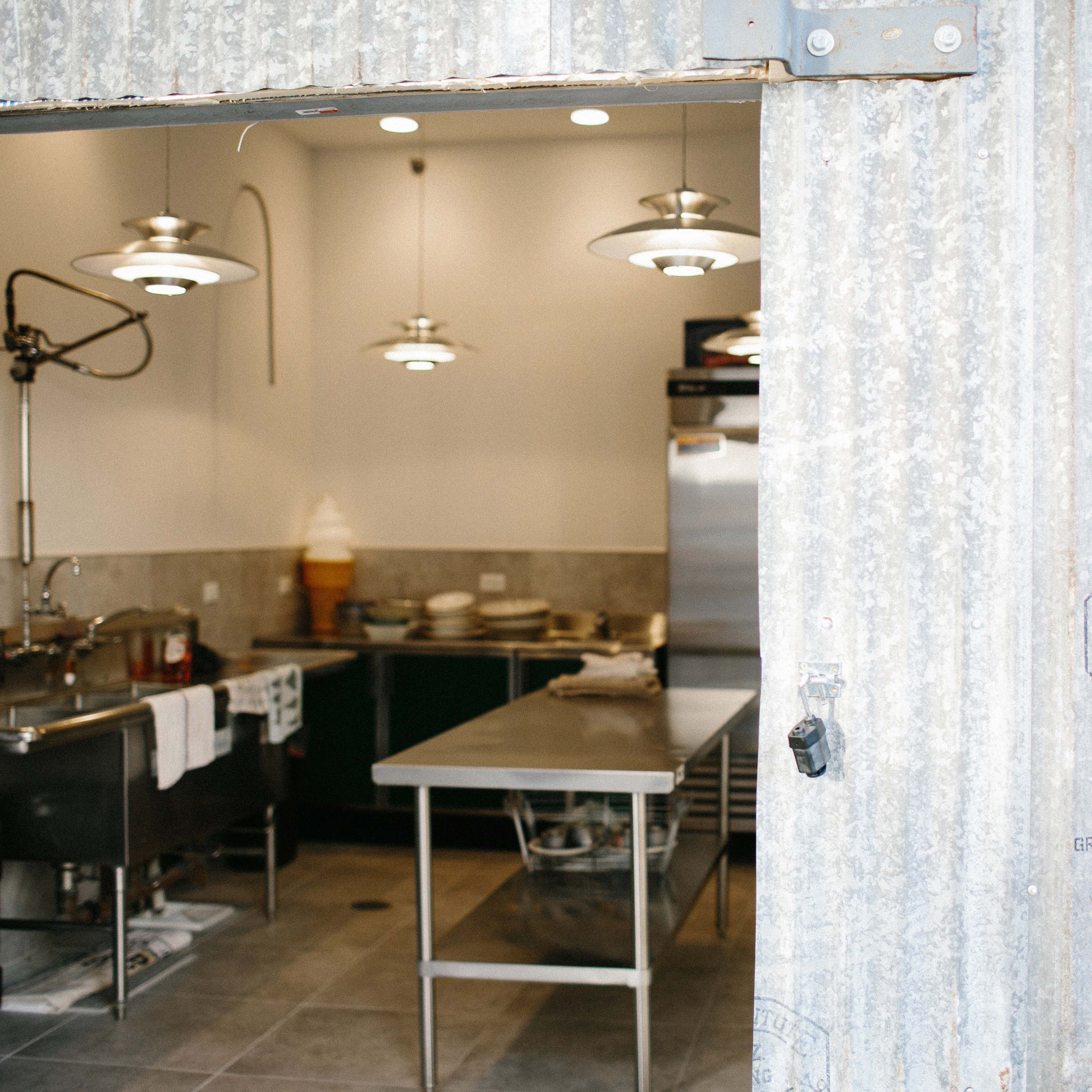 A commercial kitchen with stainless steel equipment and counters visible through a corrugated metal doorframe.