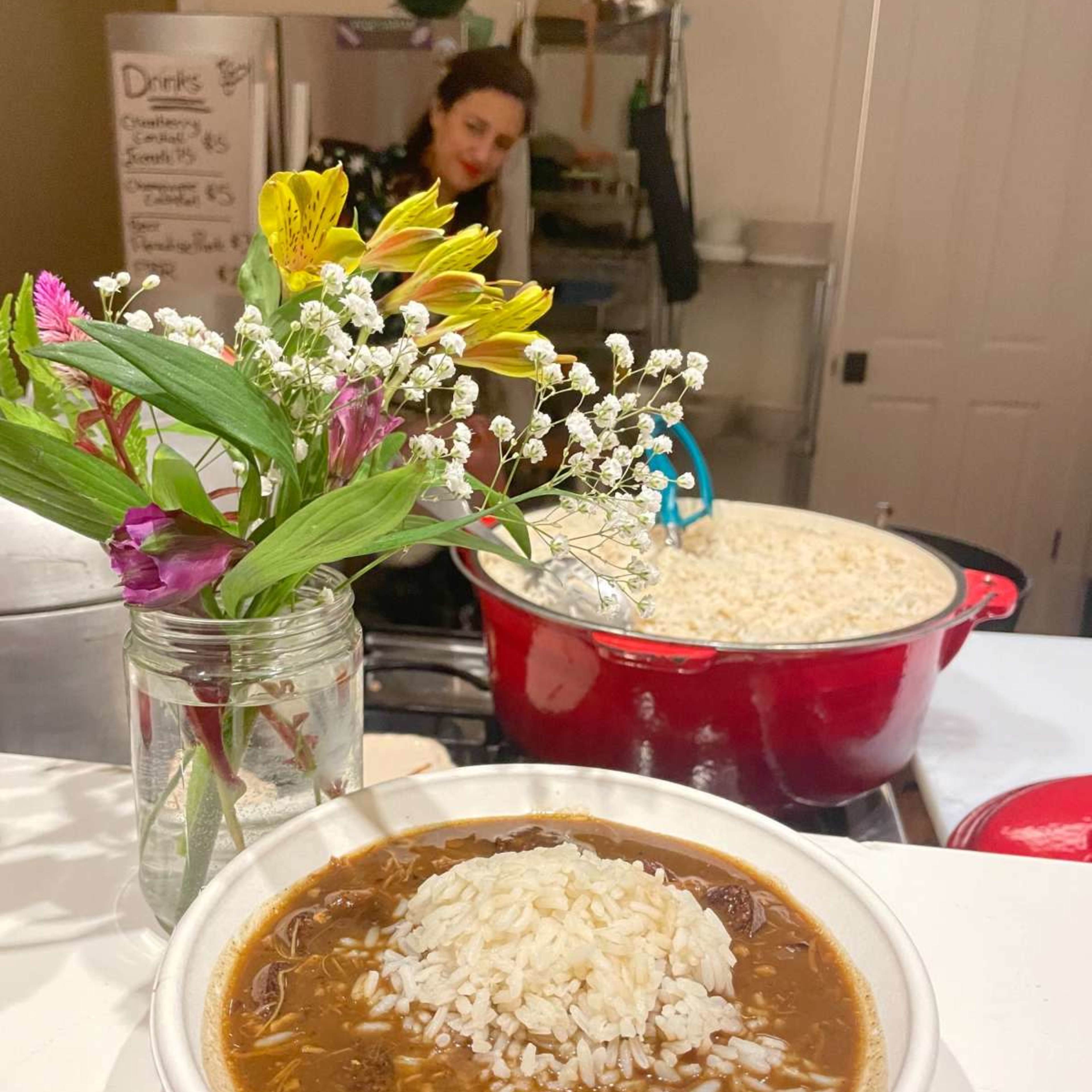 A bowl of gumbo topped with rice sits on a counter next to a vase of flowers, while a woman prepares food in the background.