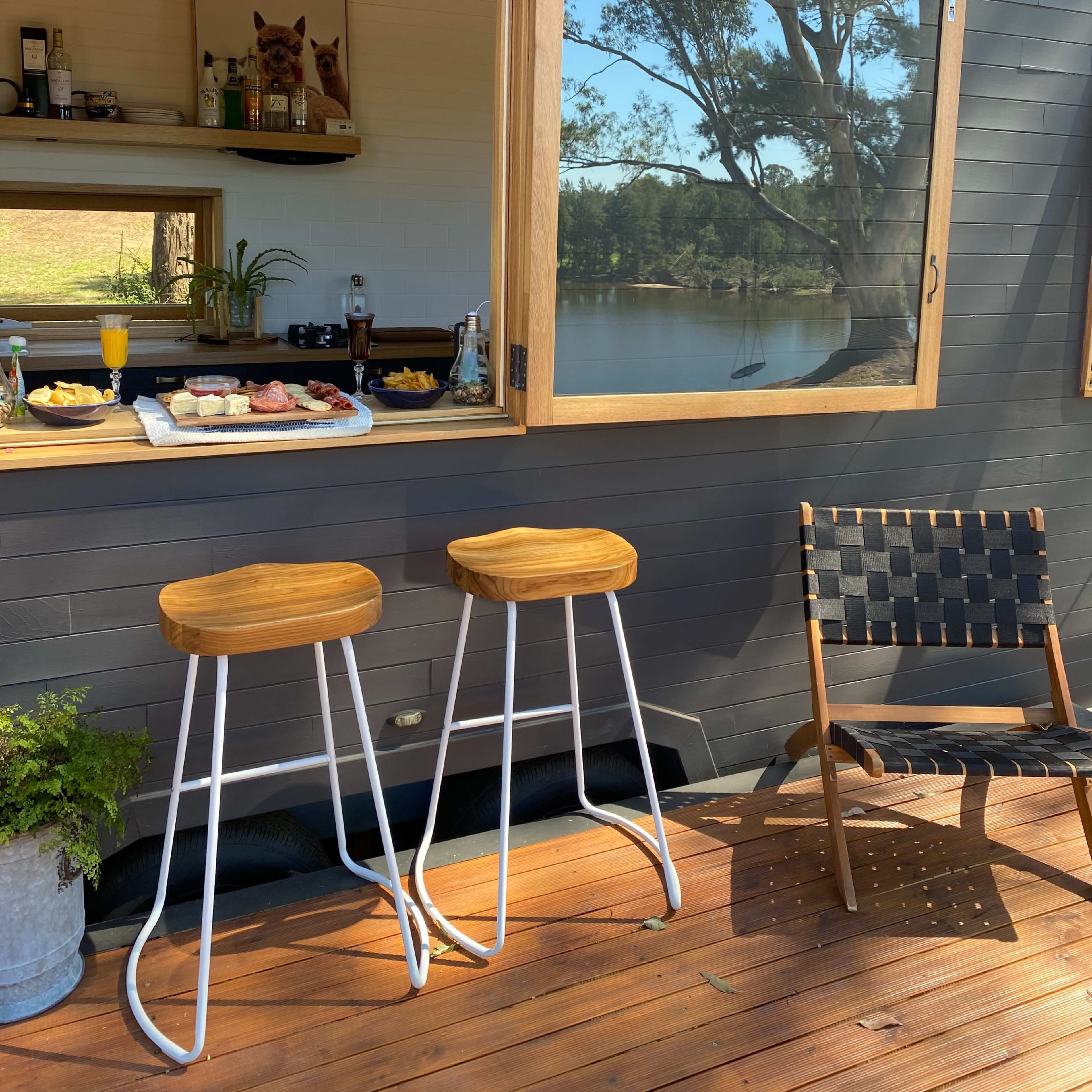 A wooden deck features two bar stools near an open window displaying a snack table and a view of a lake.