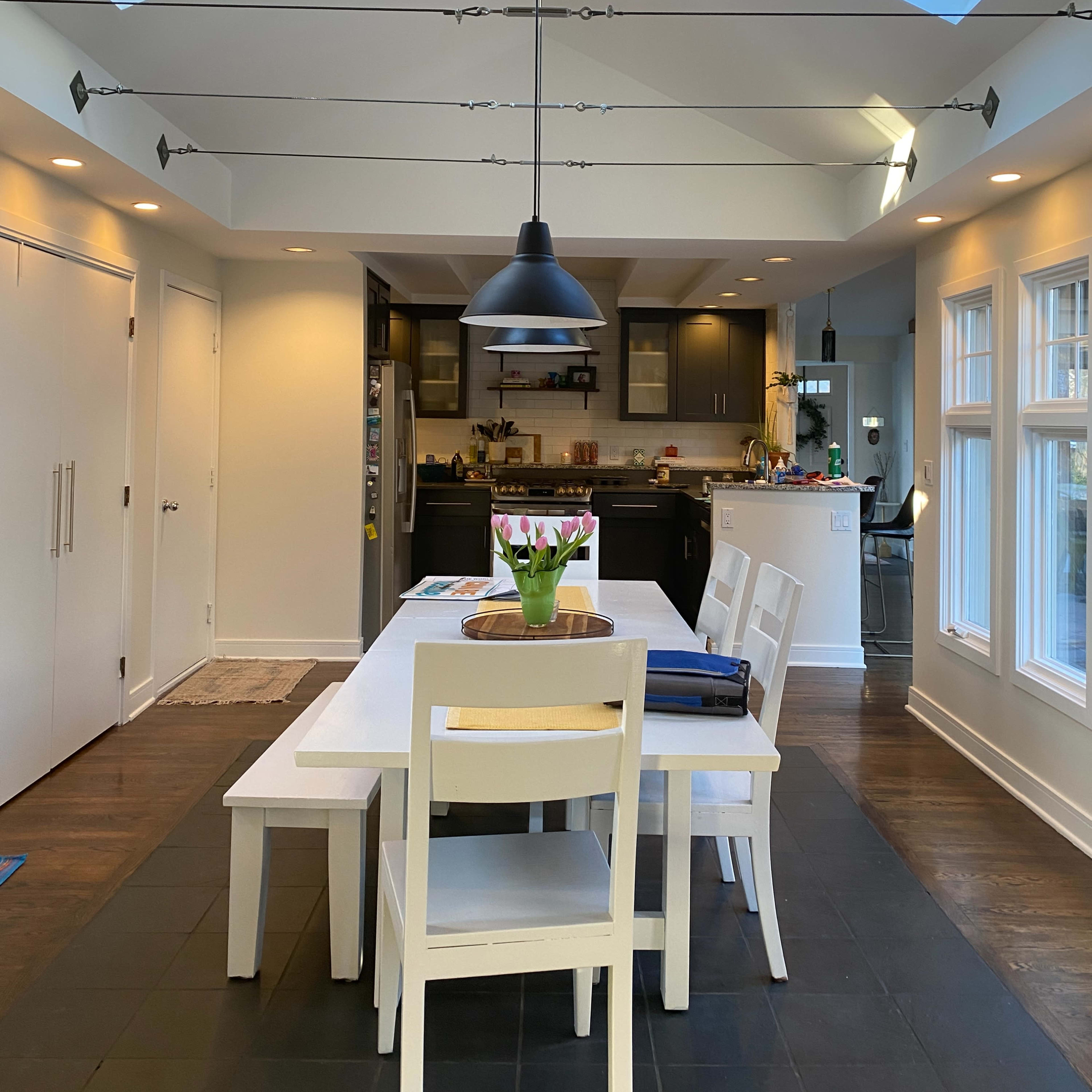 A modern kitchen and dining area featuring a white table surrounded by chairs, with skylights above and a dark-colored kitchen in the background.