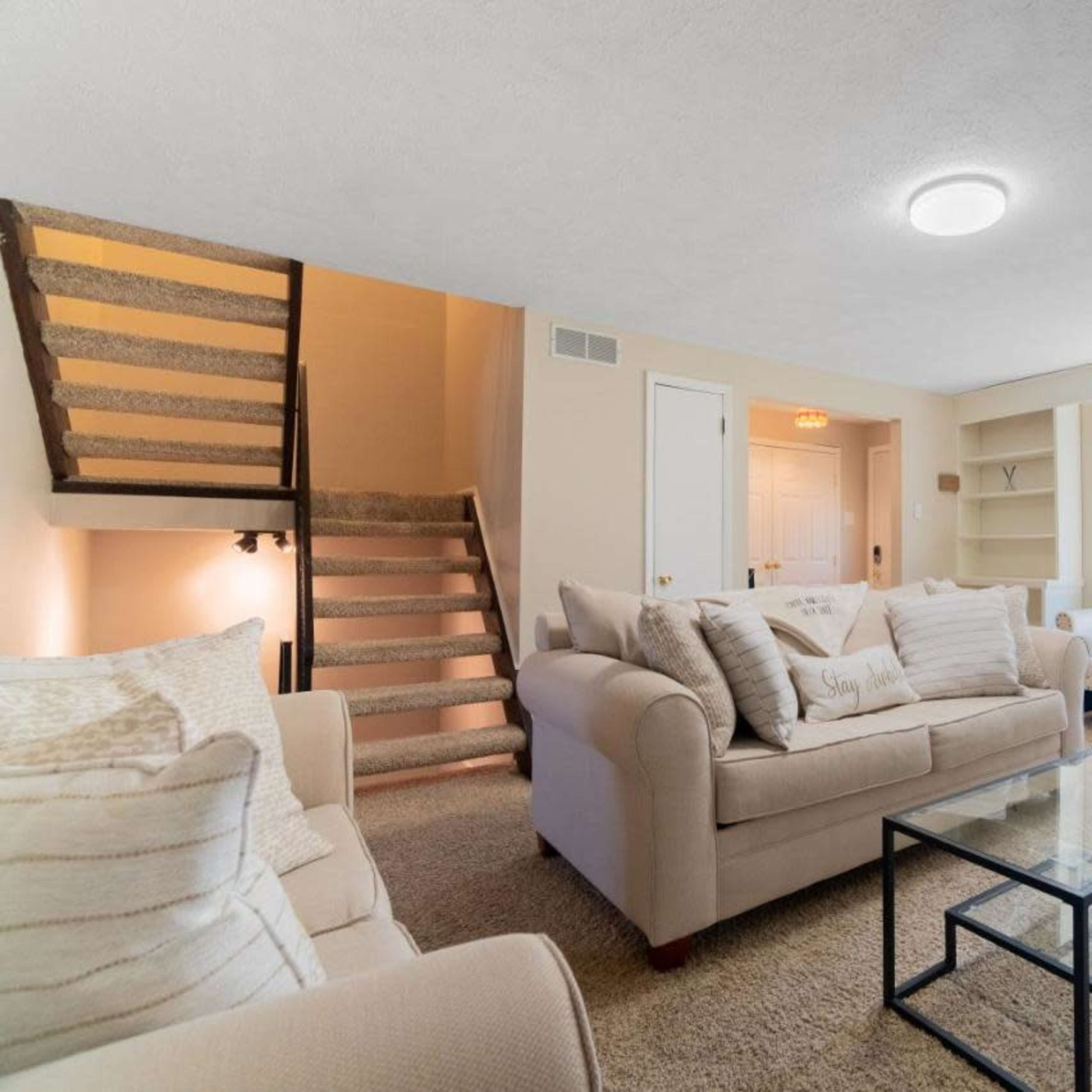 A bright living room features beige sofas, a glass coffee table, and a large window overlooking the outdoor space, with stairs leading to a second level.