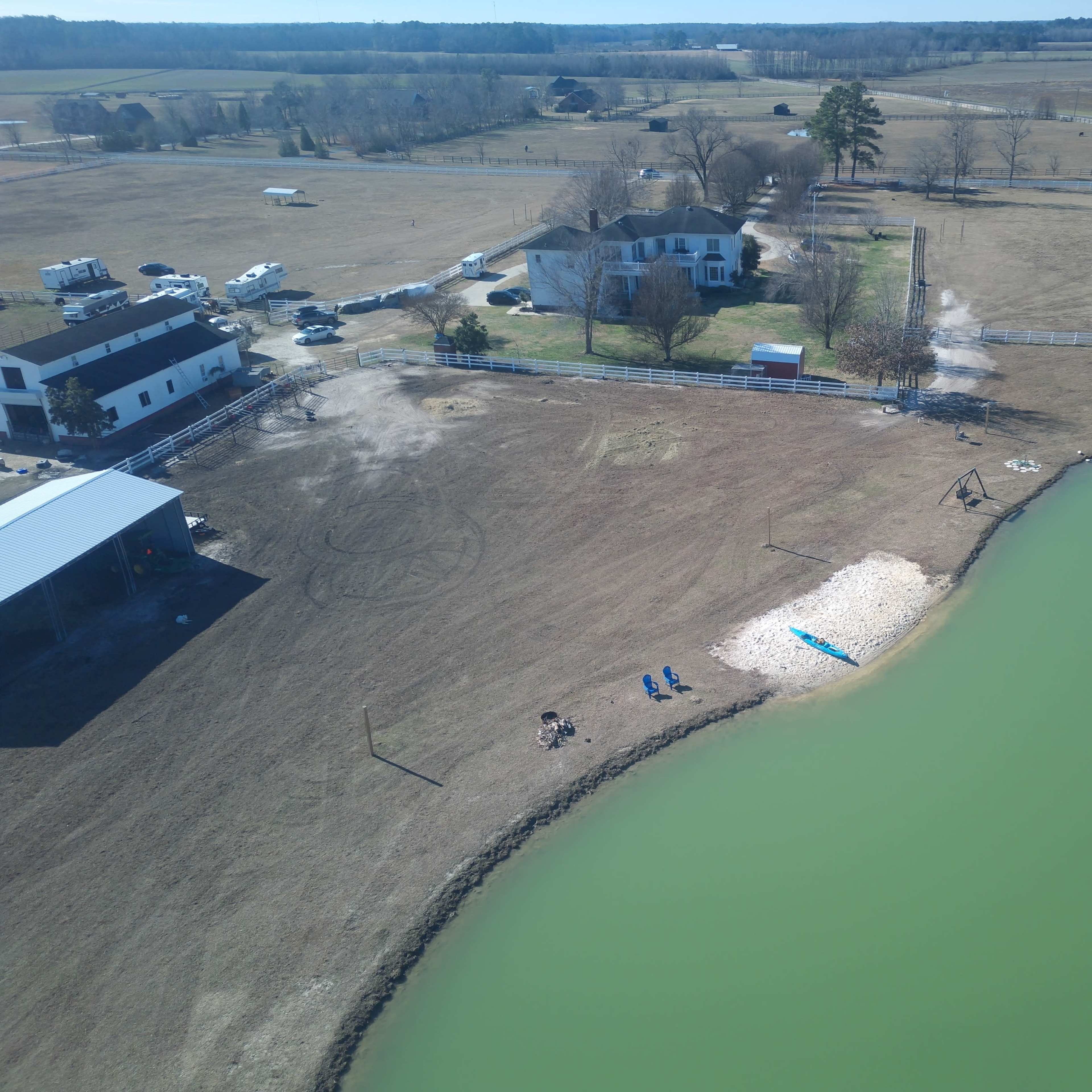 A rural landscape featuring a large house near a green pond, surrounded by open fields, trailers, and other structures.