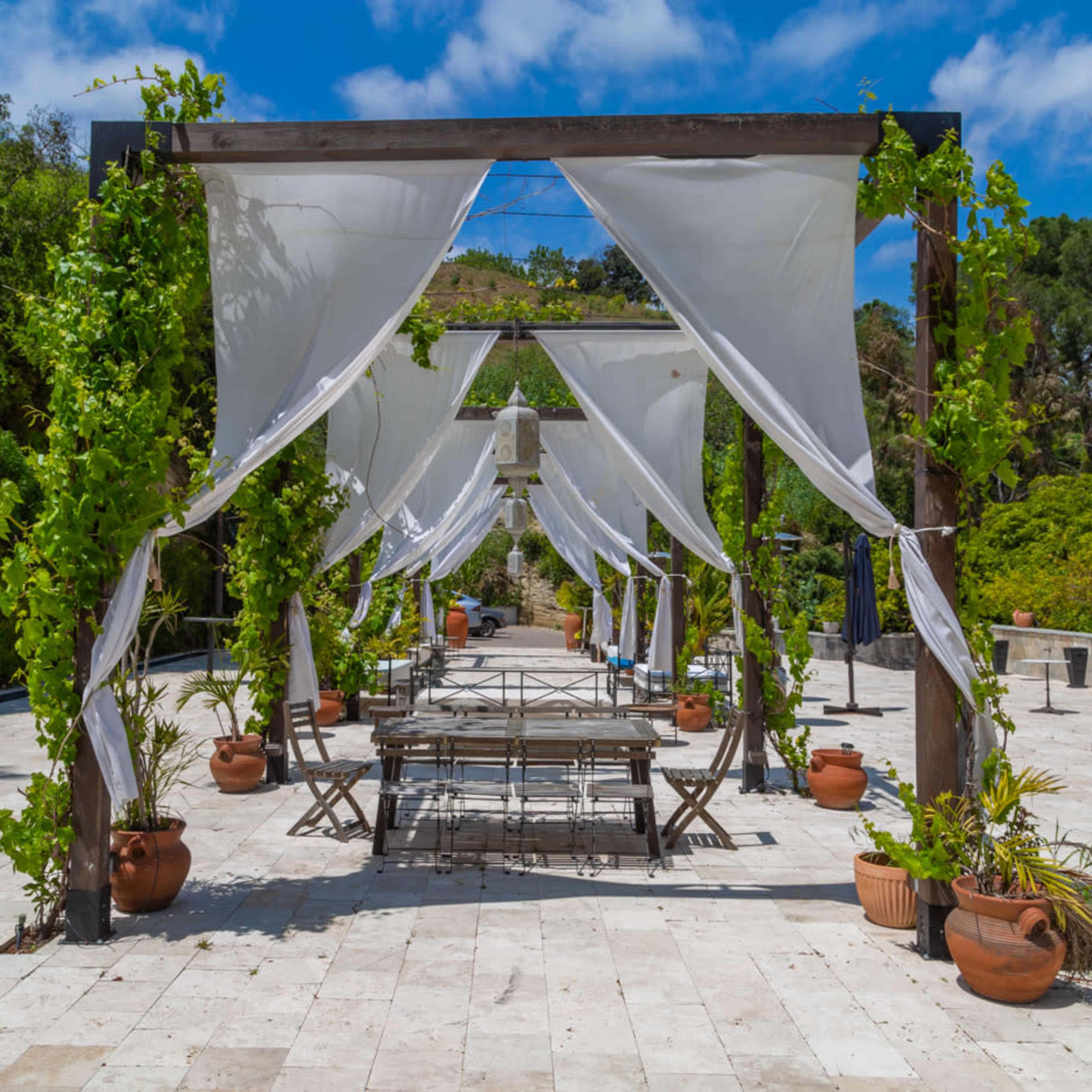 An open-air event space features a series of wooden structures draped with white fabric, flanked by potted plants along a stone pathway.
