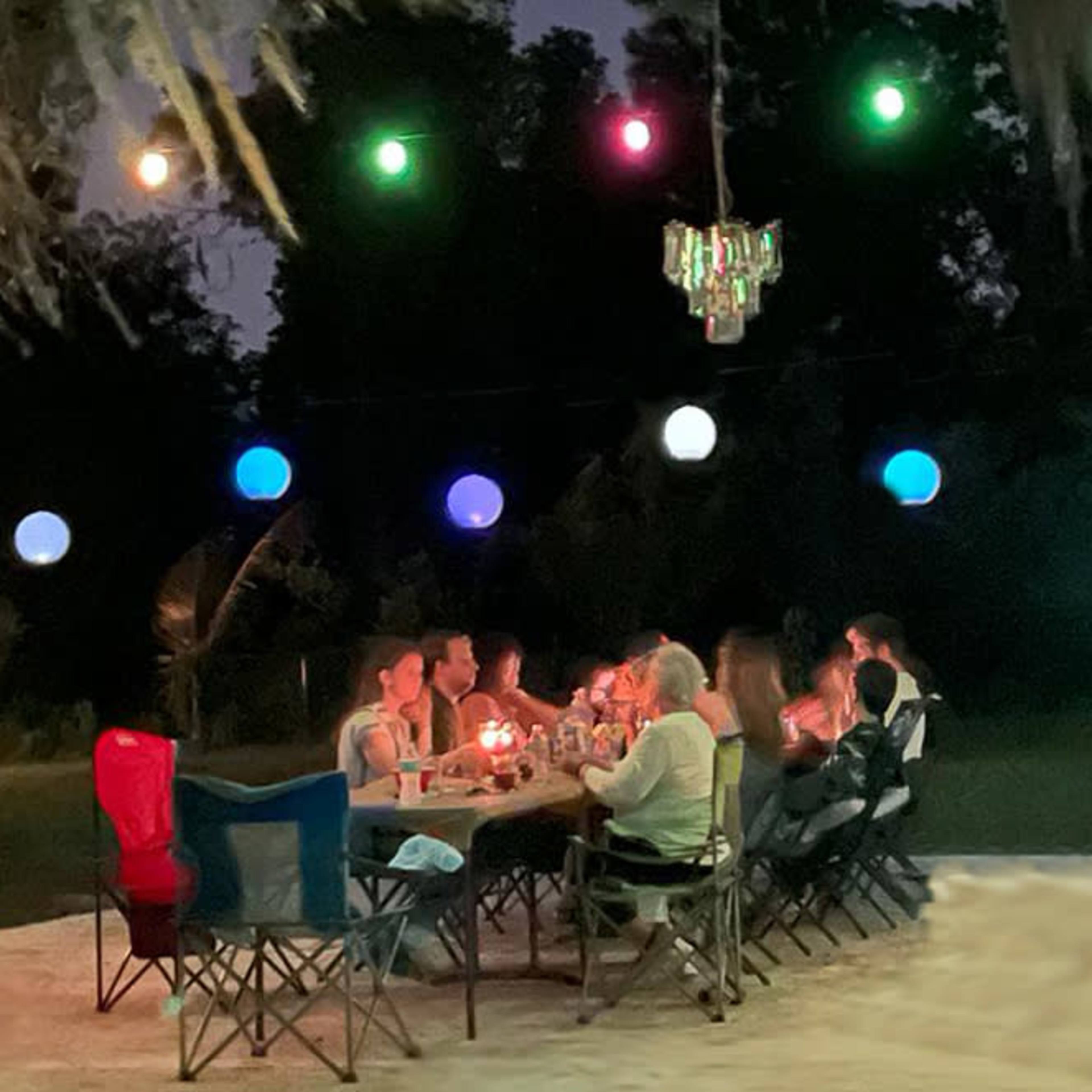 A group of people sits around a table with candles on a well-lit patio at night, surrounded by colorful hanging lanterns.