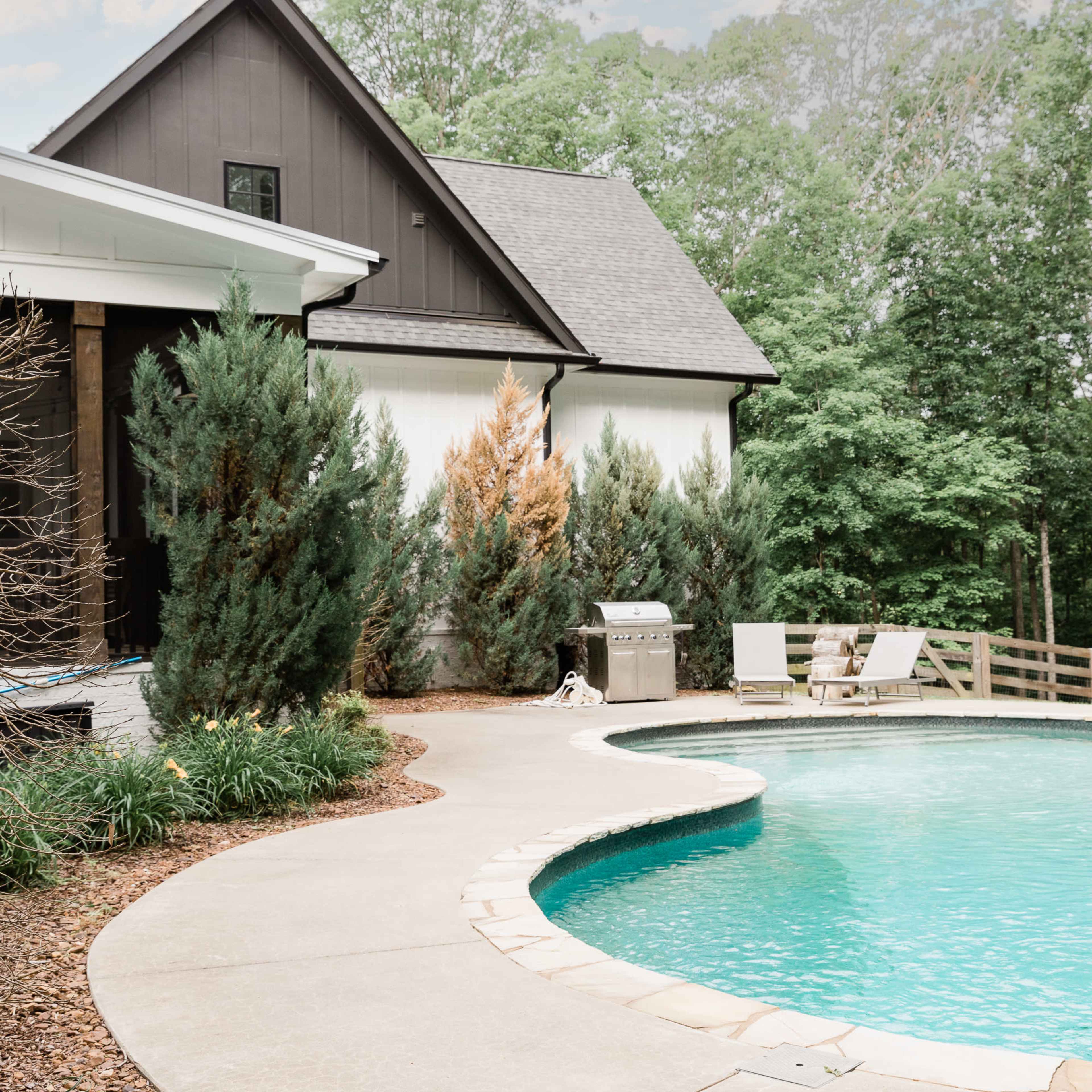 A curved concrete patio surrounds a swimming pool next to a modern house with a dark exterior and lush greenery.