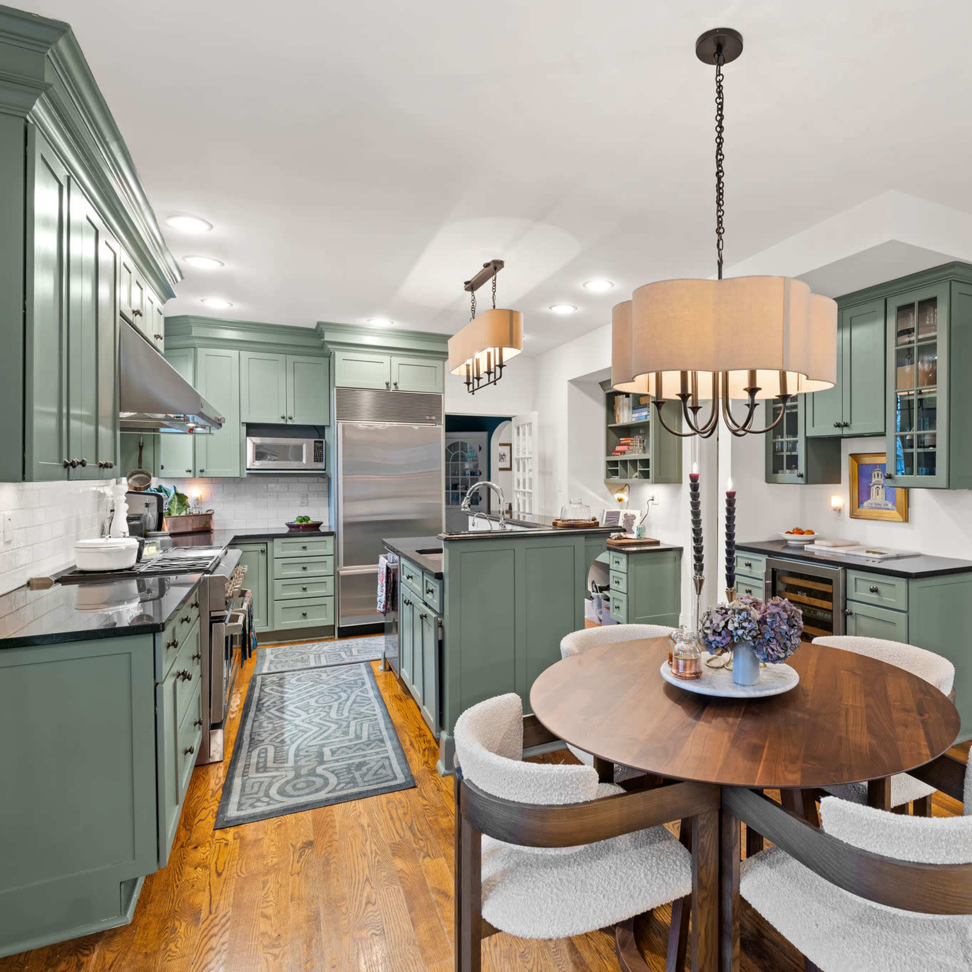 A modern kitchen features green cabinetry, stainless steel appliances, and a round wooden dining table surrounded by upholstered chairs.