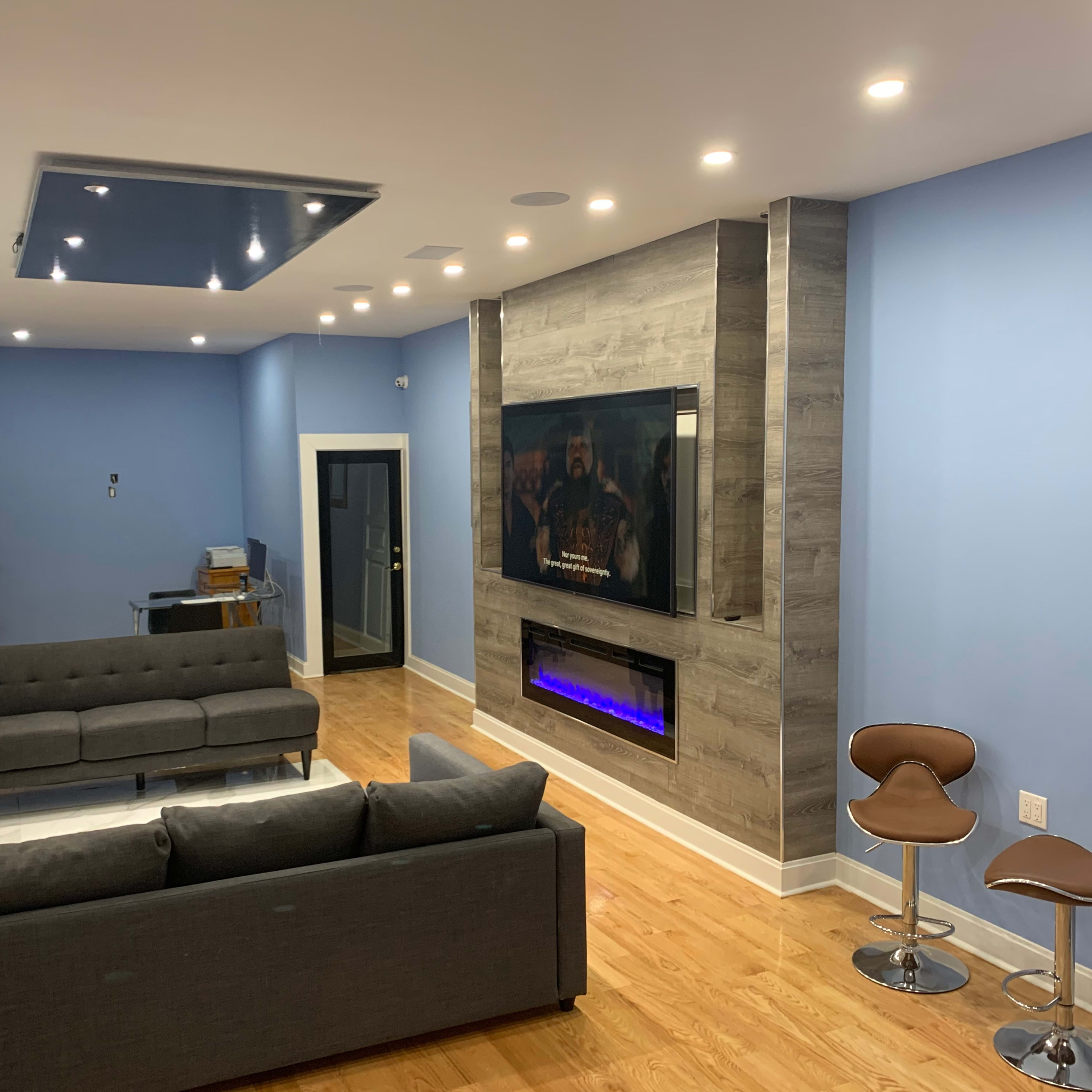 The living room features two gray sofas facing a wall-mounted television with an electric fireplace below, accompanied by modern bar stools on one side.