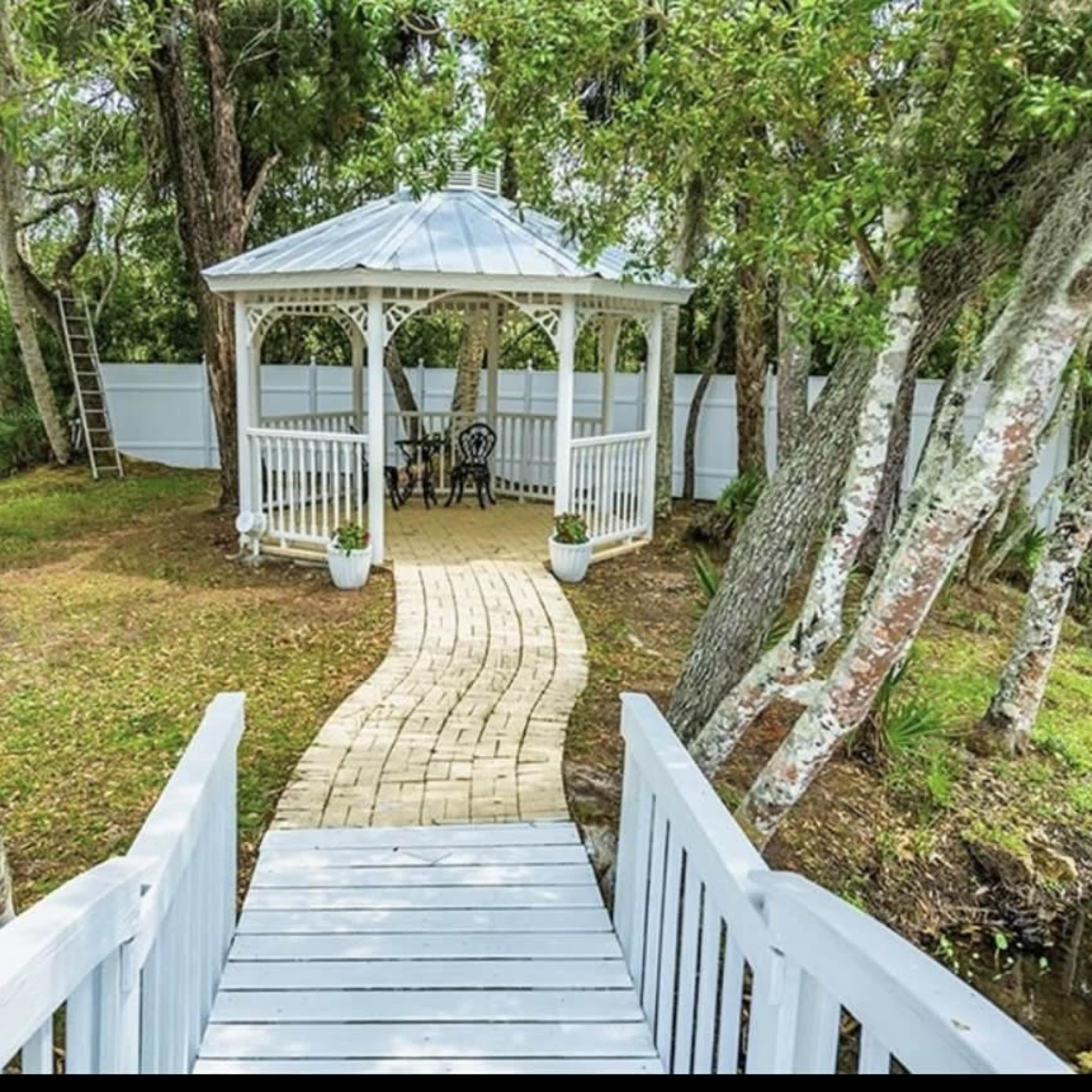 A wooden pathway leads to a white gazebo surrounded by trees in a tranquil garden setting.