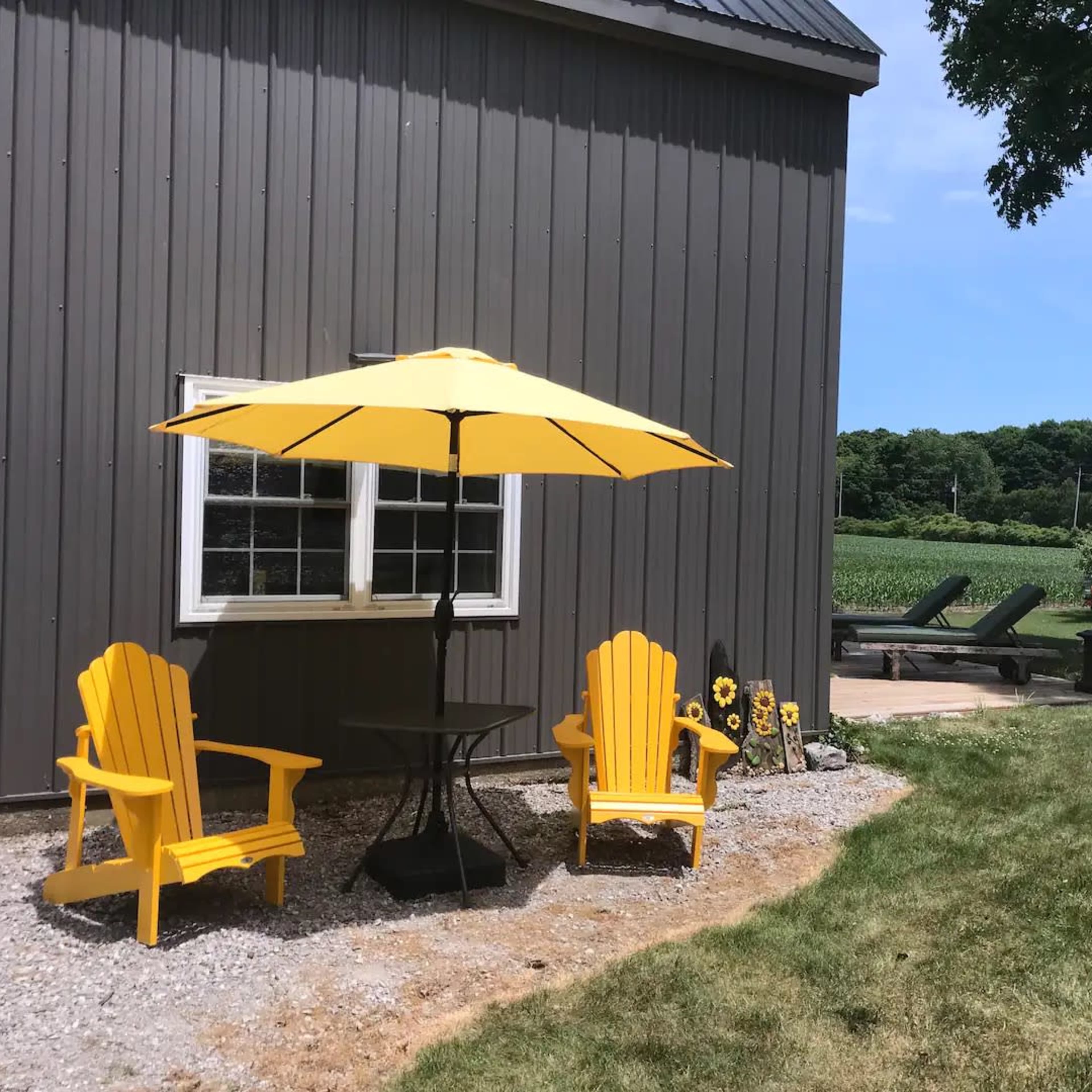 Two yellow Adirondack chairs sit beside a black table under a matching umbrella, next to a well-manicured lawn and a field in the background.