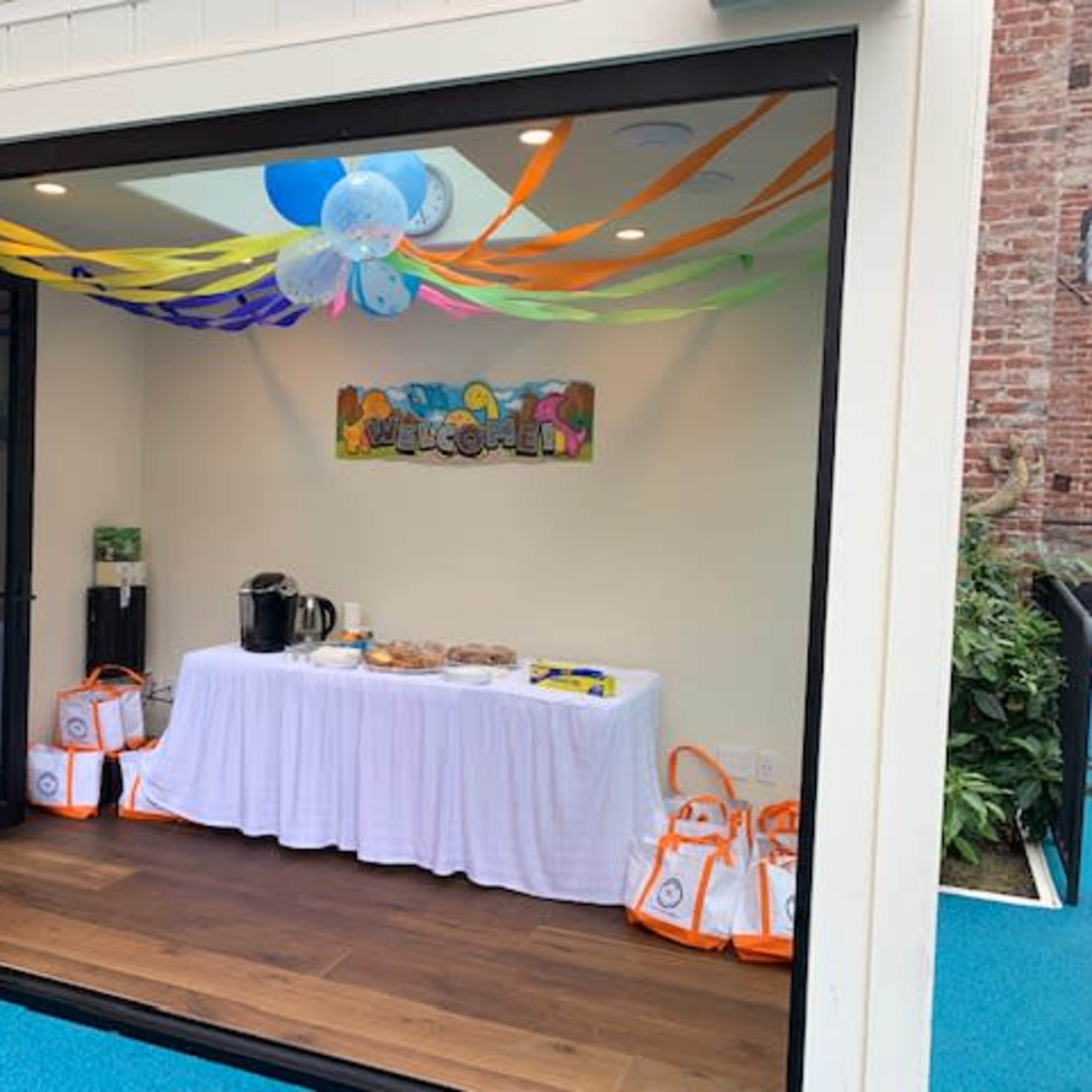 A decorated room with a table covered in a white cloth displaying snacks and drinks, adorned with colorful streamers and balloons.