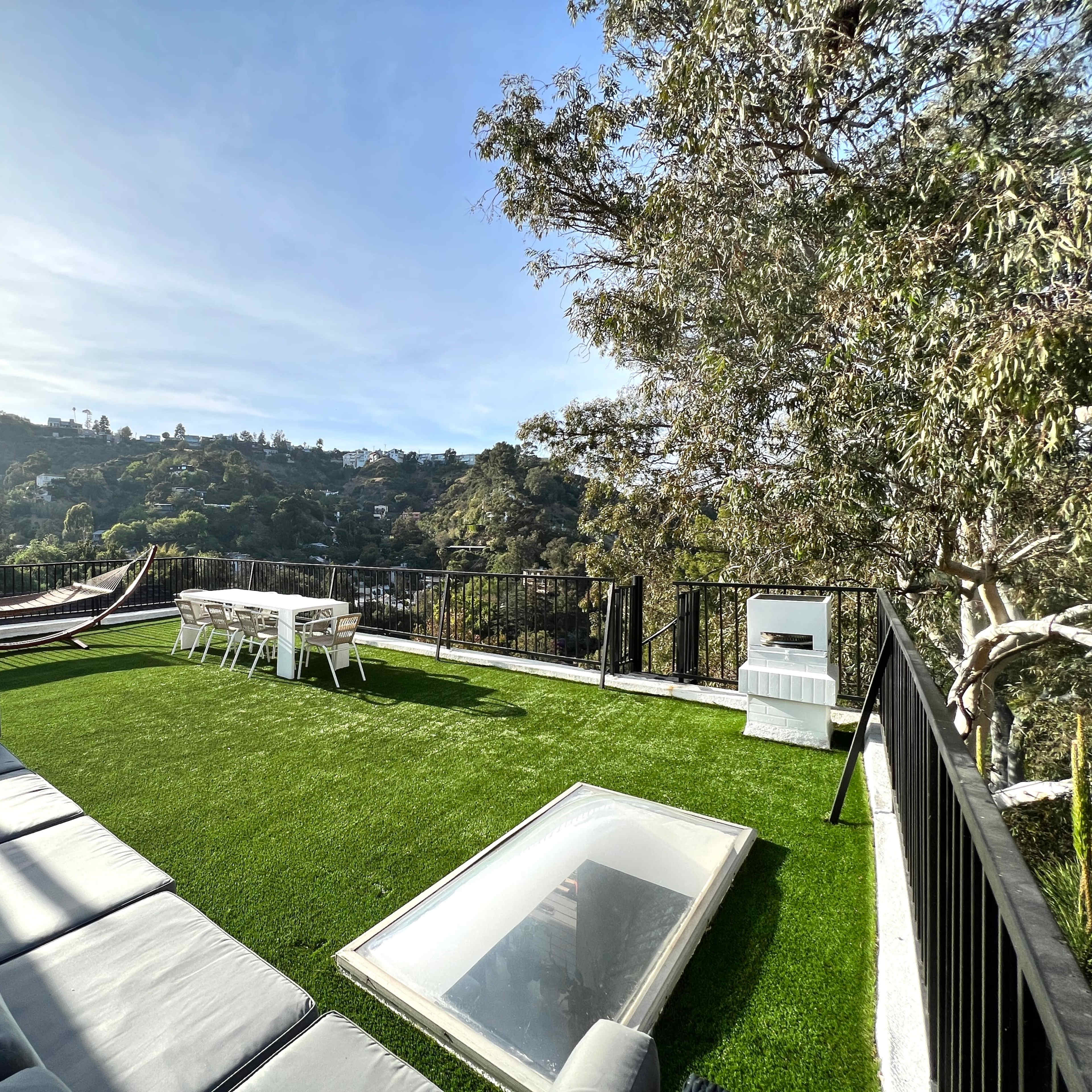 A rooftop patio with a green lawn, a hammock, a dining table, and a sectional sofa overlooking a hilly landscape.