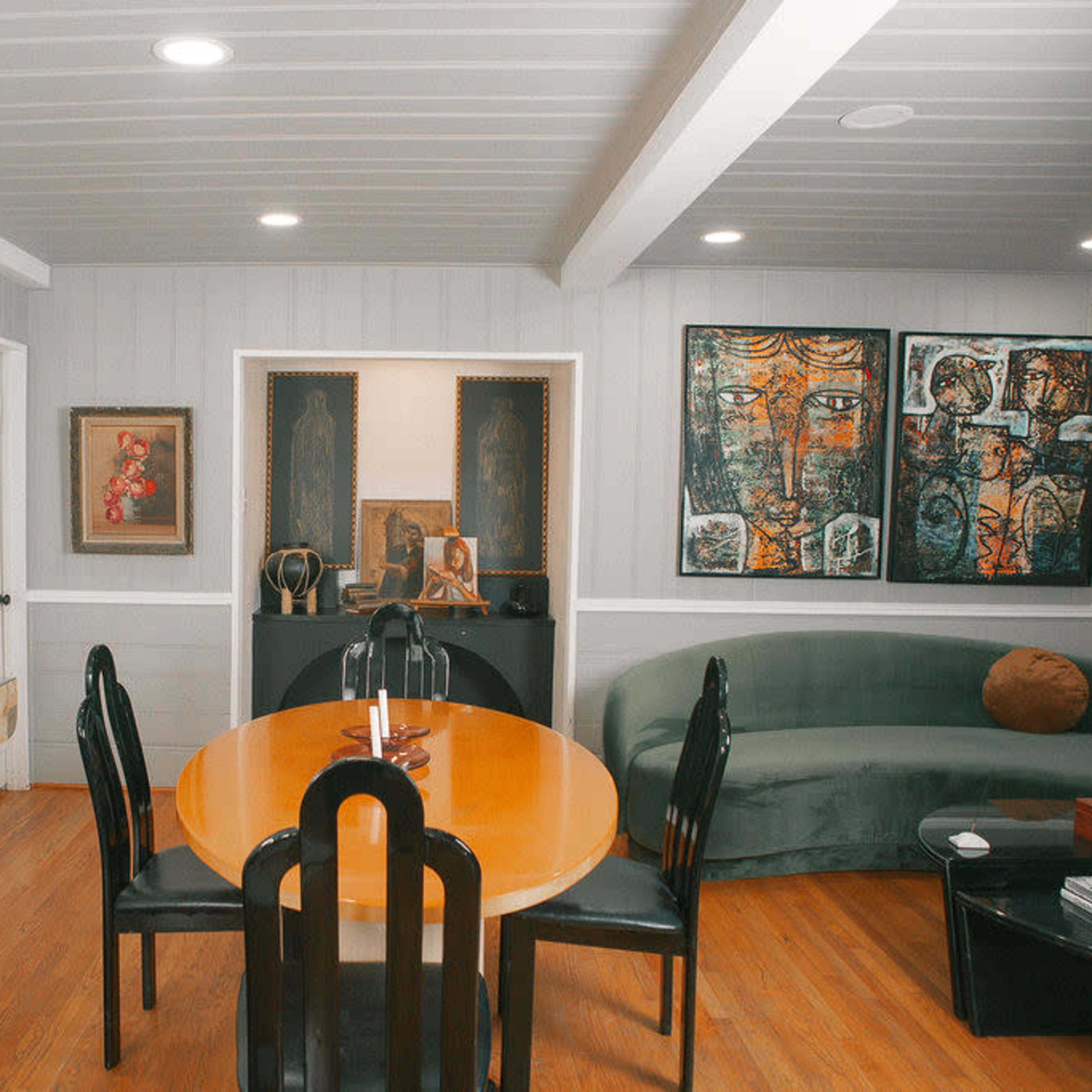 A modern living space featuring a round orange dining table, a green couch, and artwork displayed on the walls.
