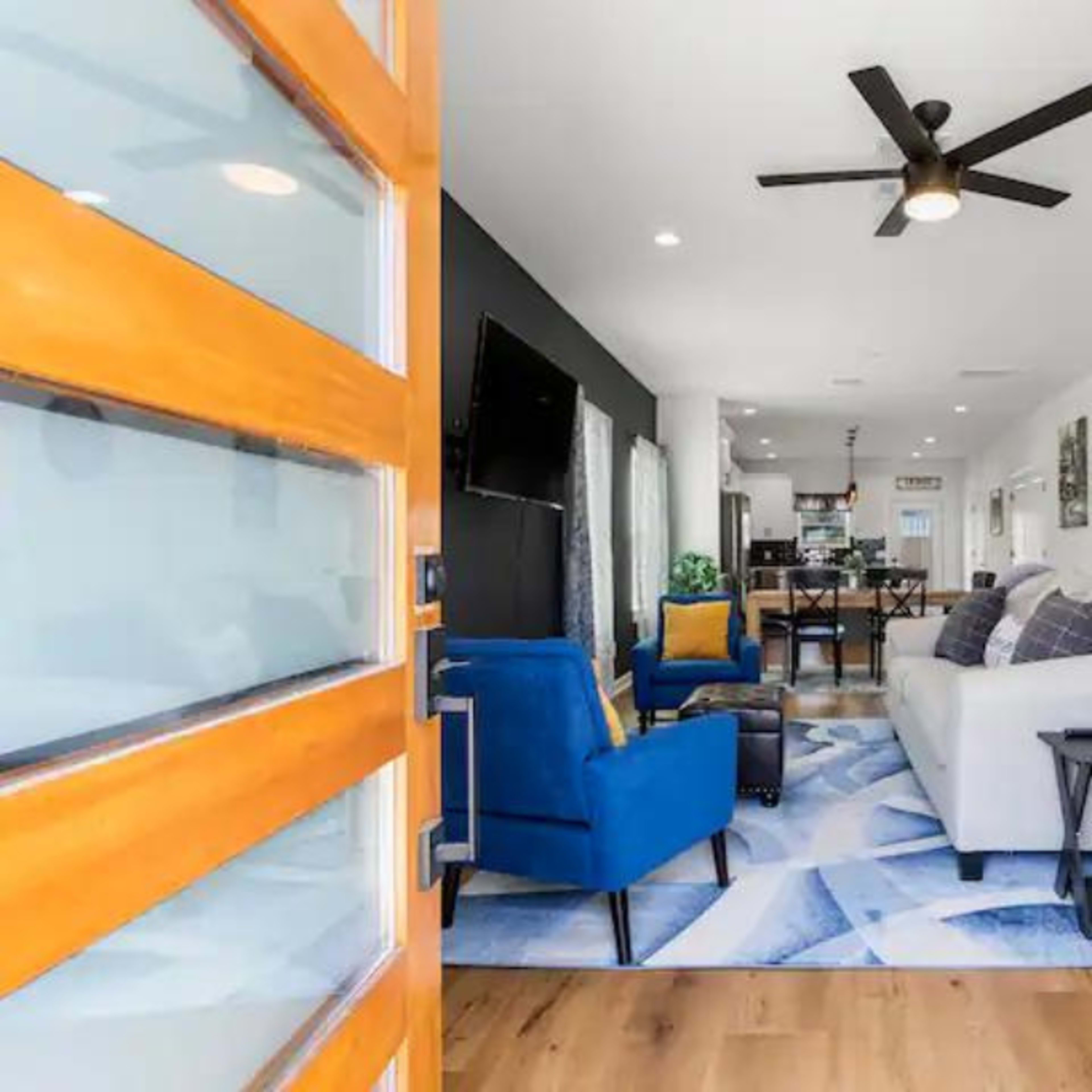 A modern living room with a blue sofa, a white couch, and a wooden door entrance leading to an open space that includes a dining area and kitchen.