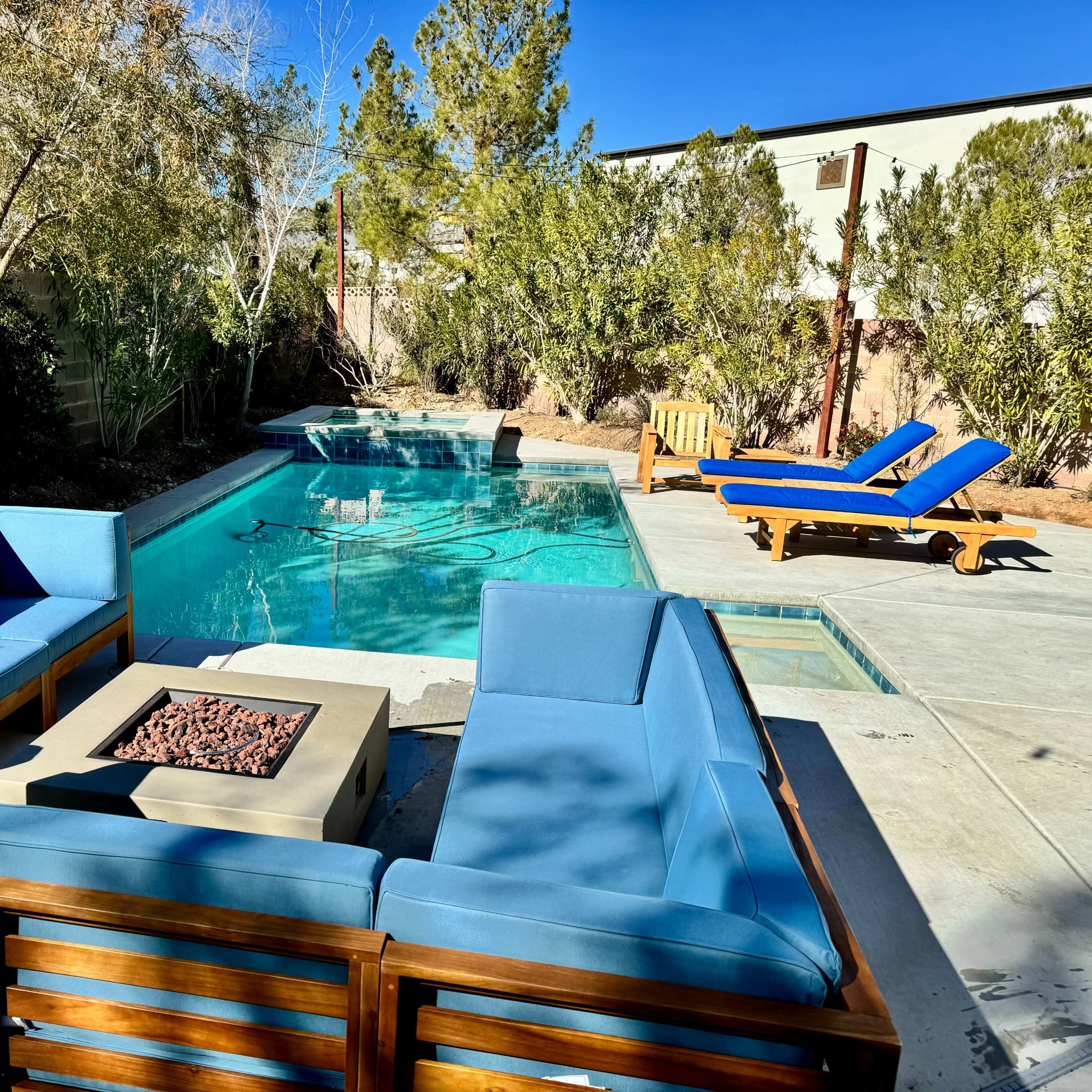 A back patio area with a seating arrangement around a fire pit, a swimming pool, and a lounge chair under clear blue skies.