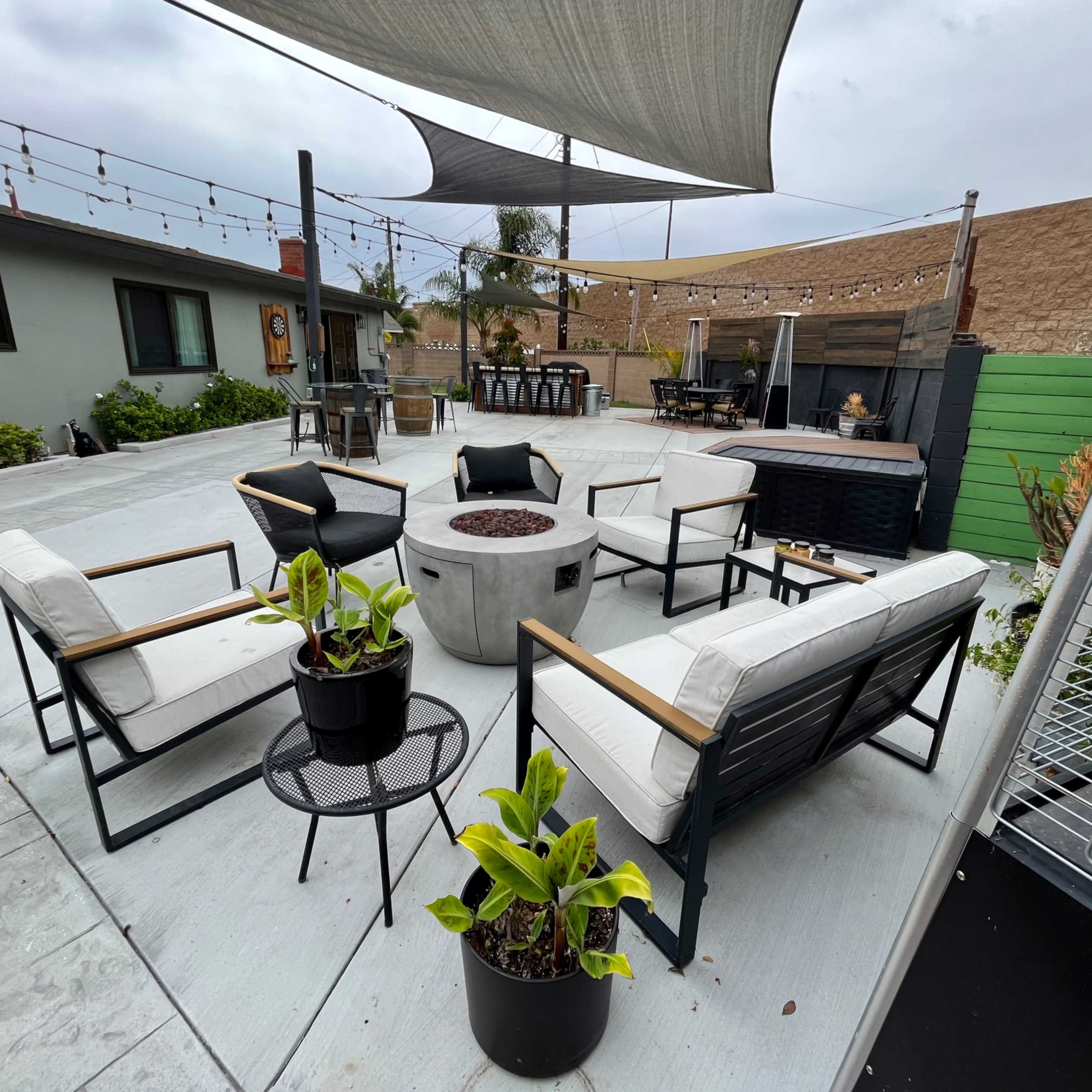 A modern outdoor seating area with several chairs, a central fire pit, and shade sails overhead, surrounded by green plants and a barbecue area in the background.