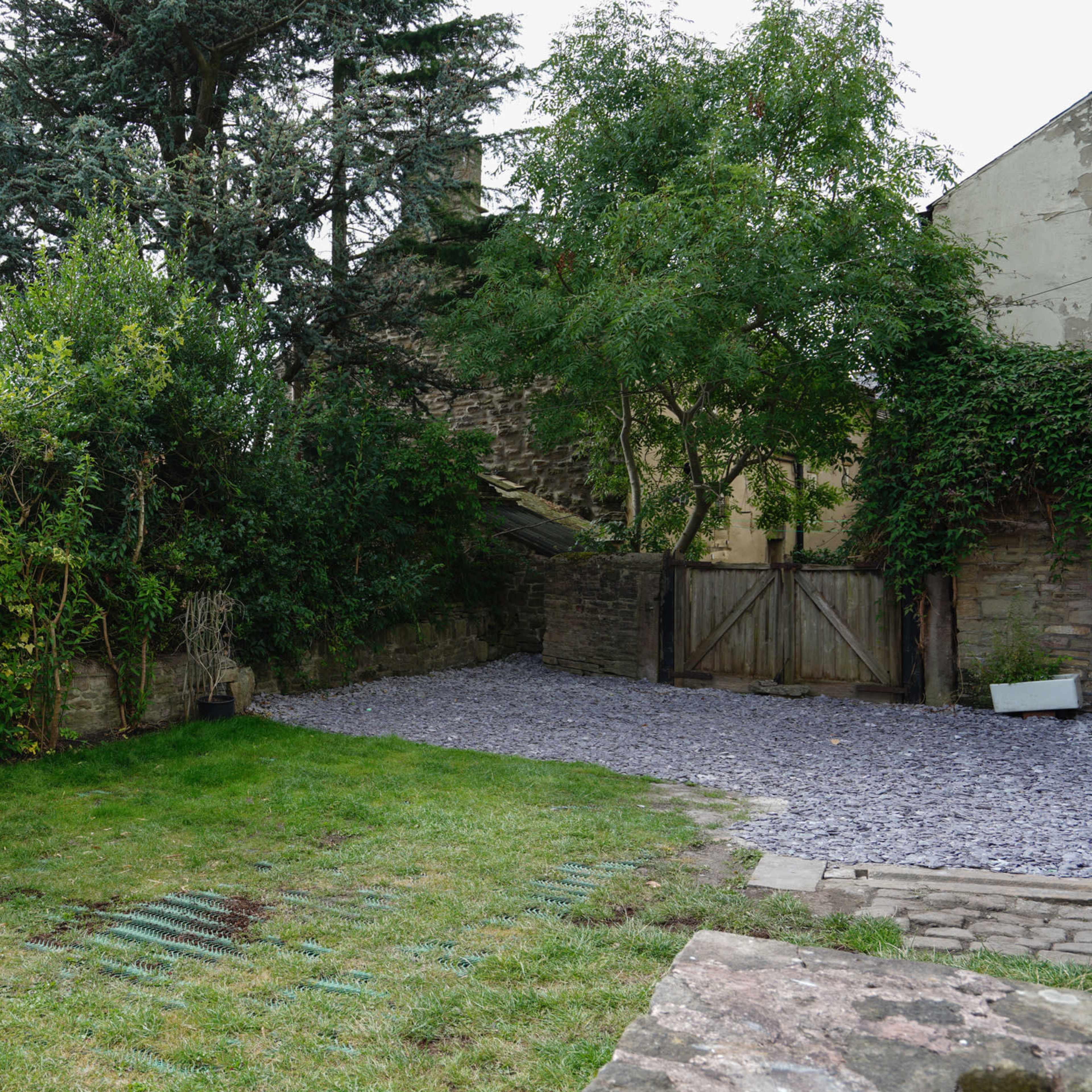 A grassy area bordered by dense greenery and a gravel-covered space, with a wooden gate leading to a background wall.