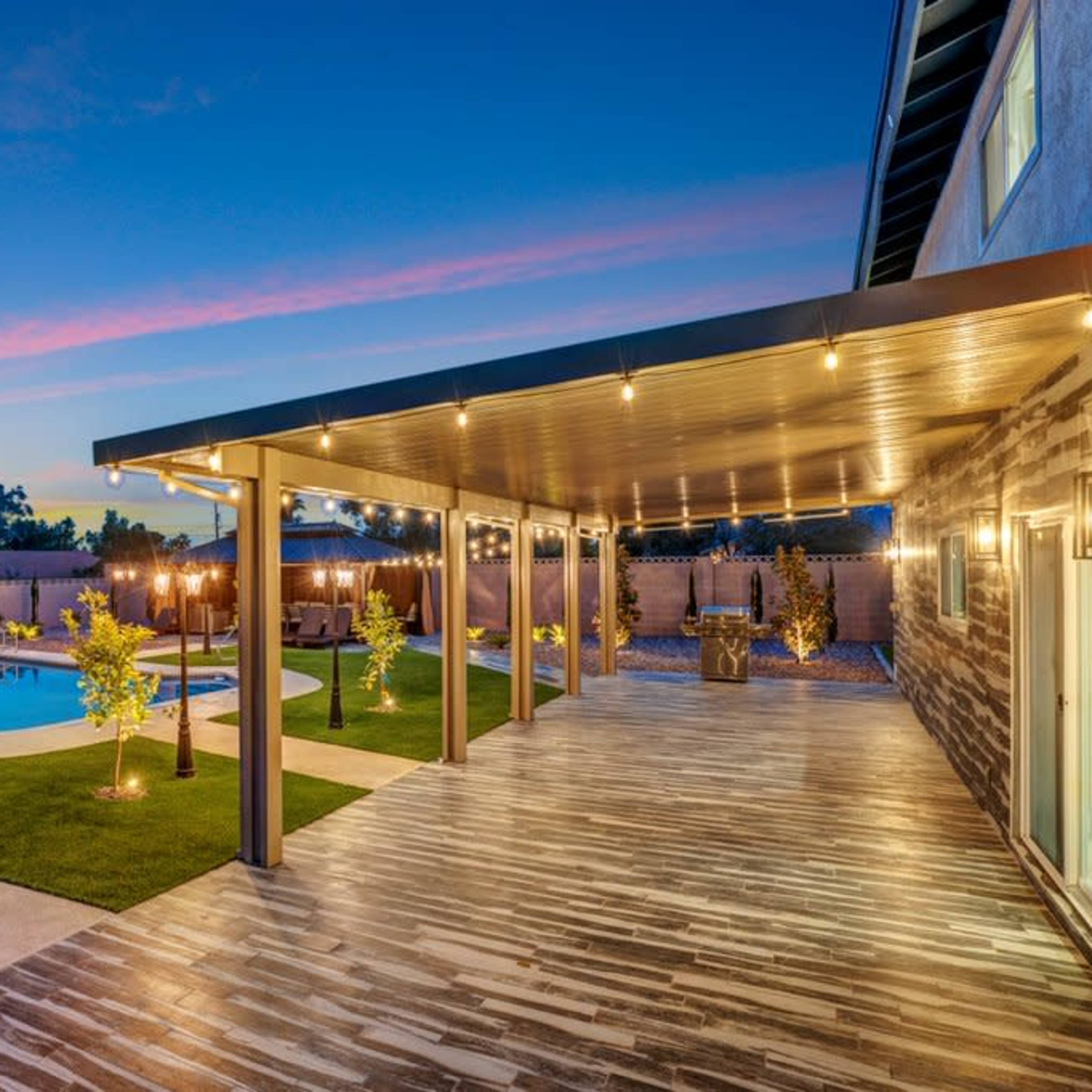 A well-lit backyard featuring a swimming pool, a covered patio area with overhead lights, and landscaped greenery under a twilight sky.