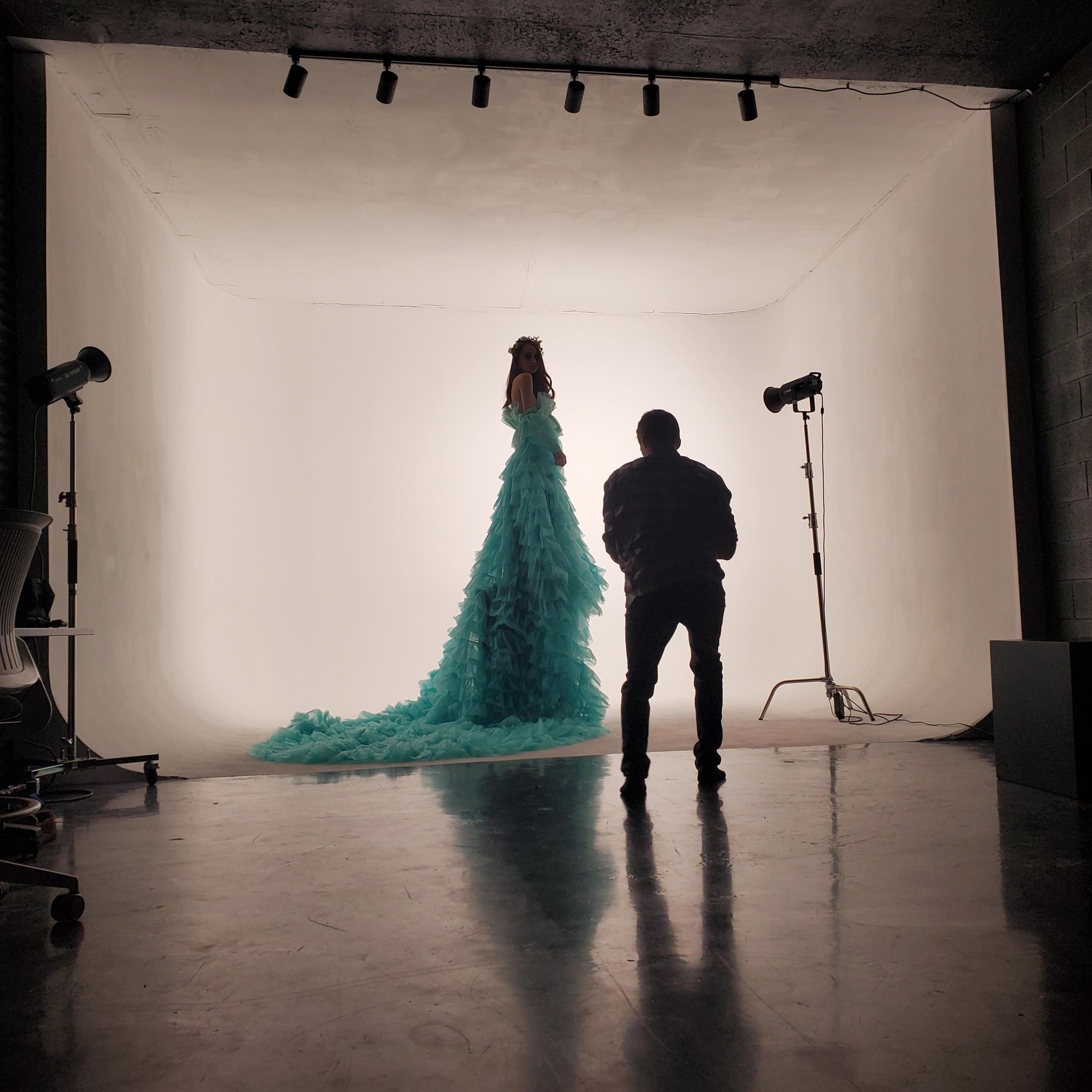 A model in a long, textured turquoise gown stands against a brightly lit backdrop while a photographer captures the scene in a studio.