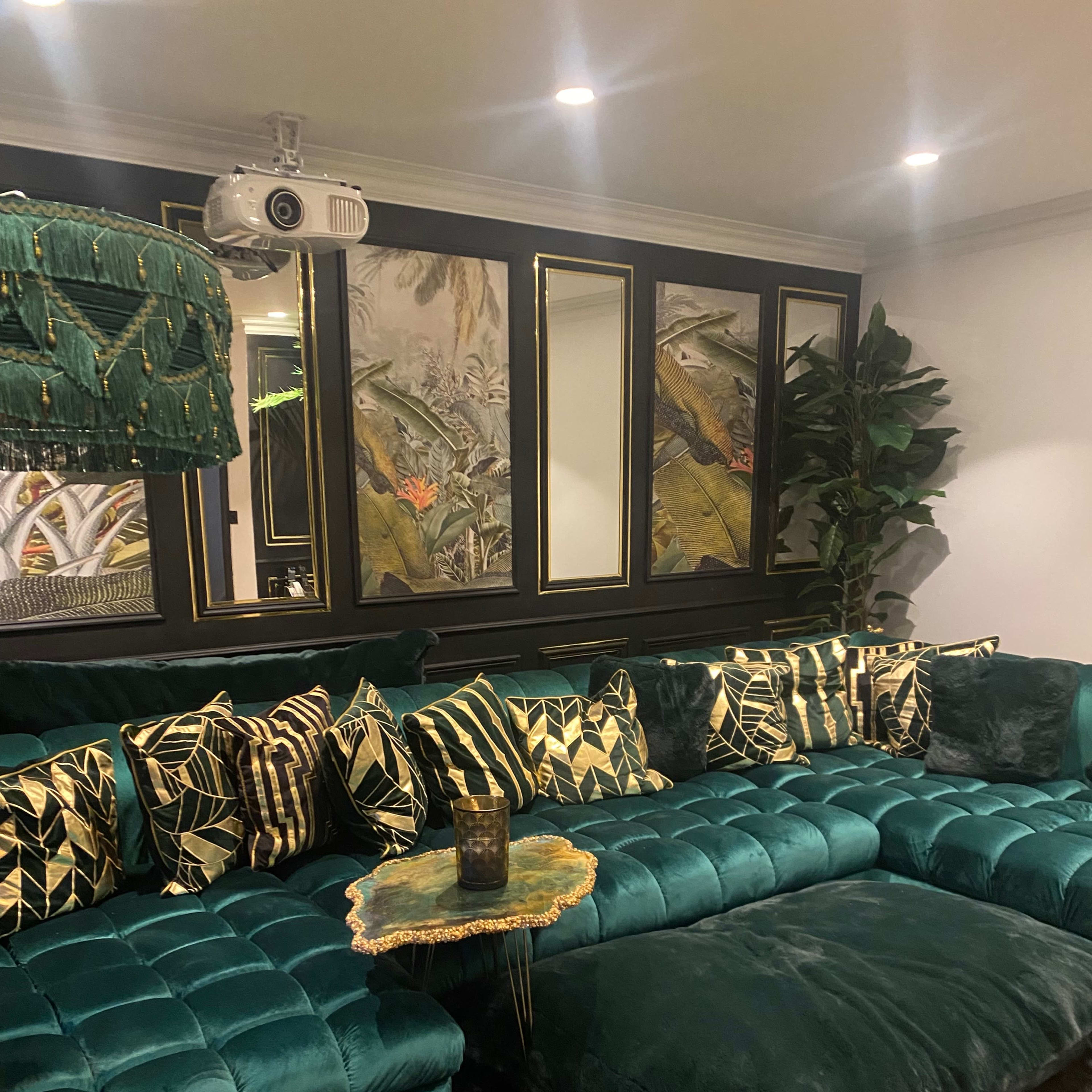 A luxurious living room featuring a green velvet sectional sofa adorned with patterned cushions, a small decorative table, and a projector mounted on the ceiling.