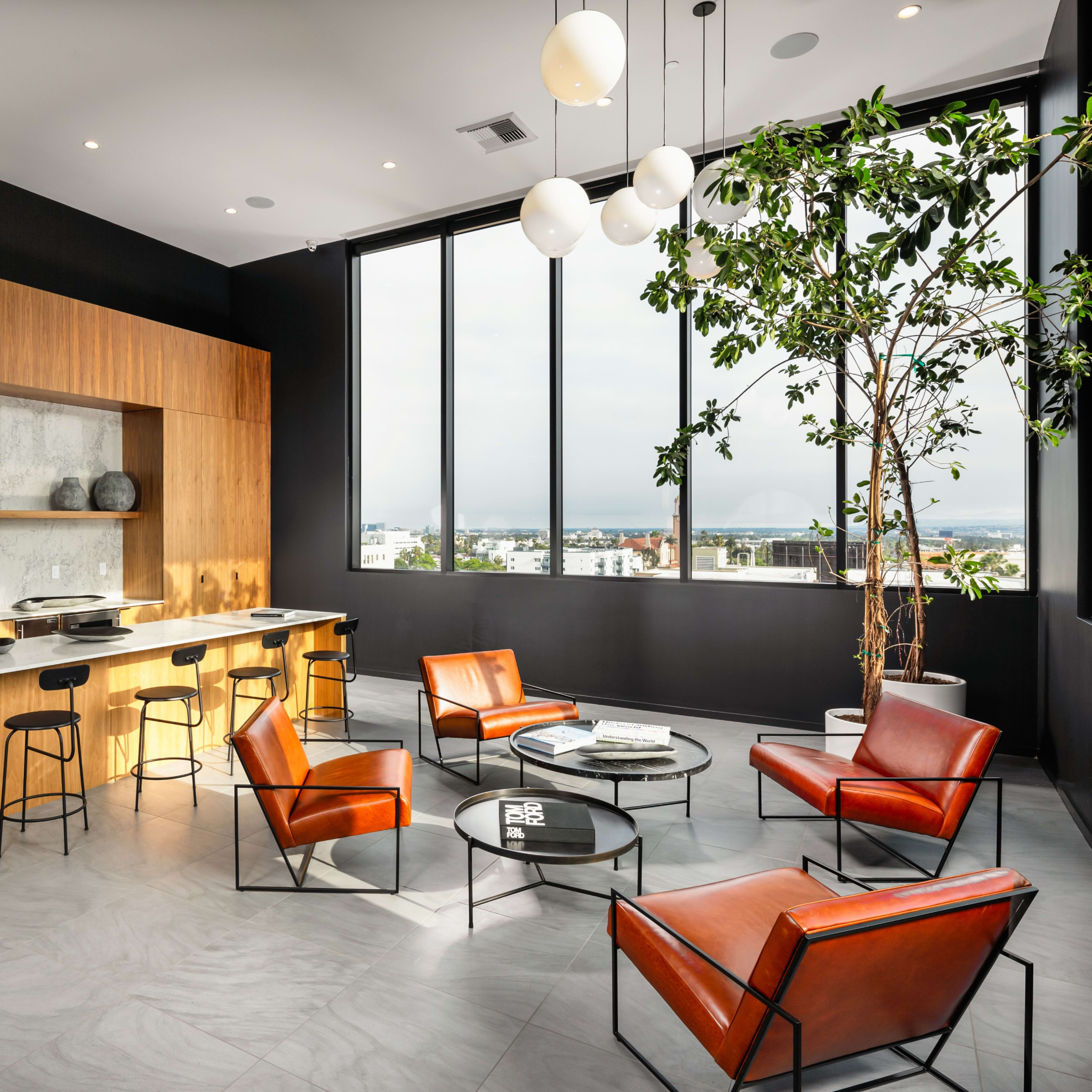 A modern lounge area features a wooden cabinetry wall, a marble countertop, four orange chairs, a round glass coffee table, and large windows displaying a cloudy view.