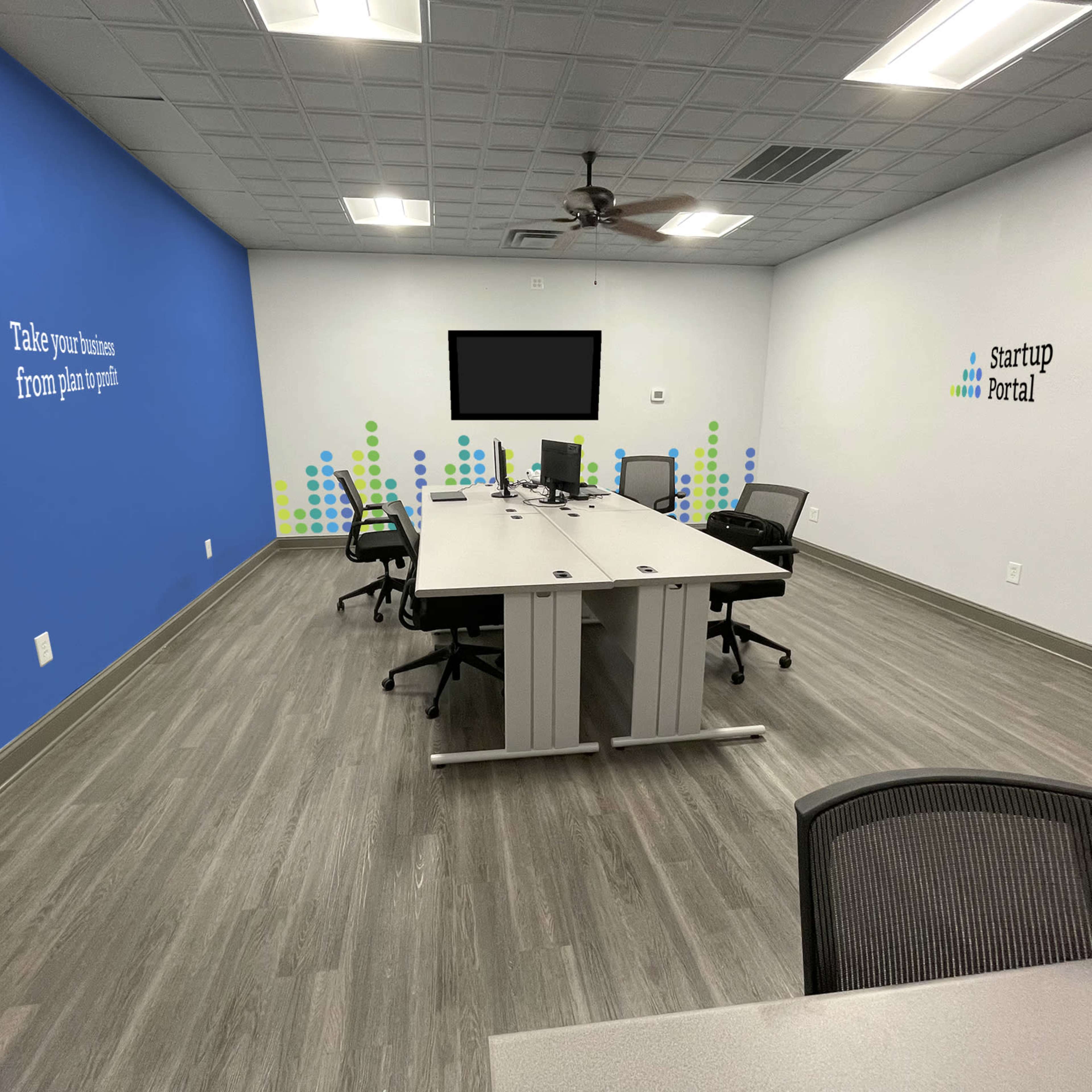 A modern, empty meeting room with a large table, four chairs, a wall-mounted television, and motivational wall graphics.