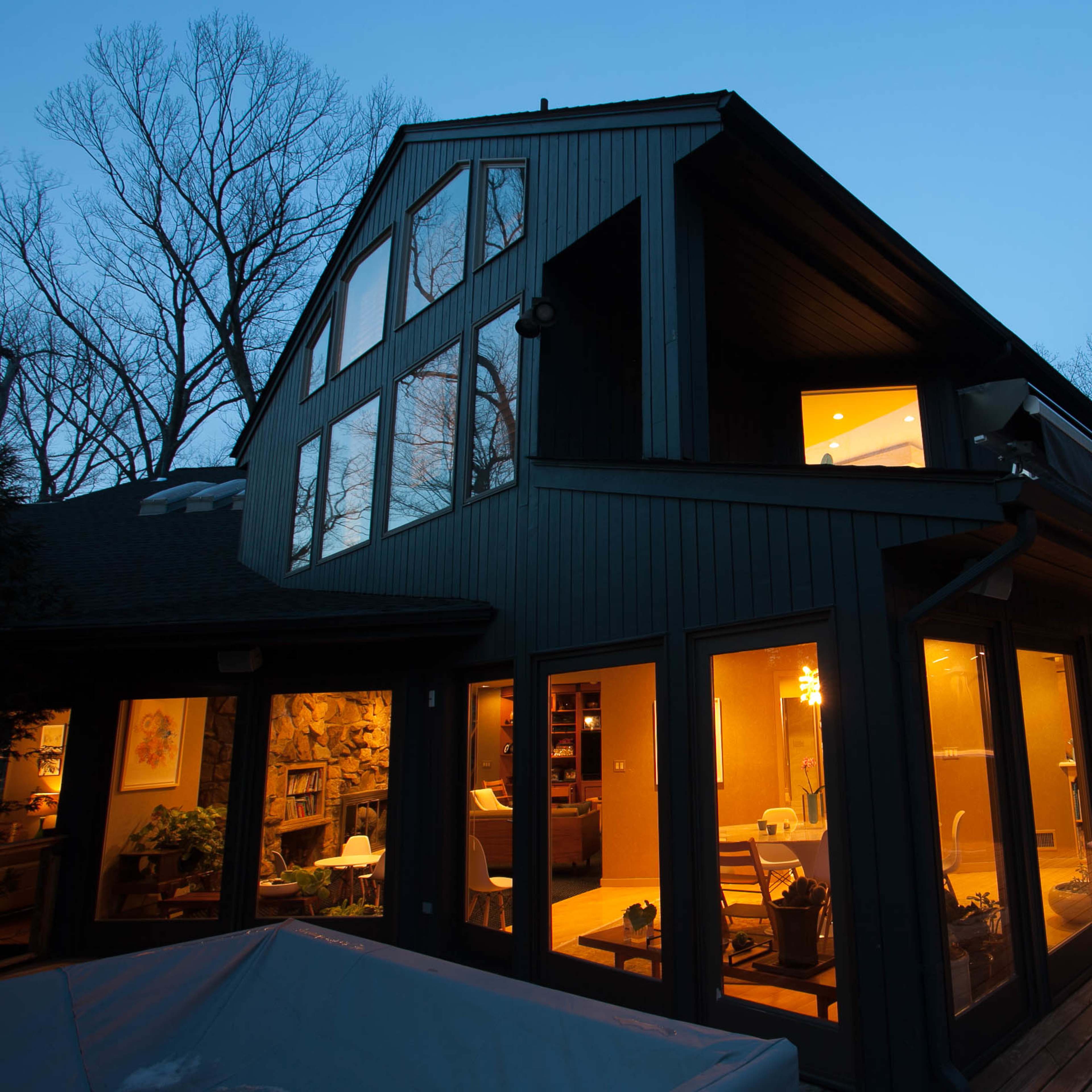 A modern, dark-colored house with large windows is partially illuminated at dusk, revealing warm light from within.