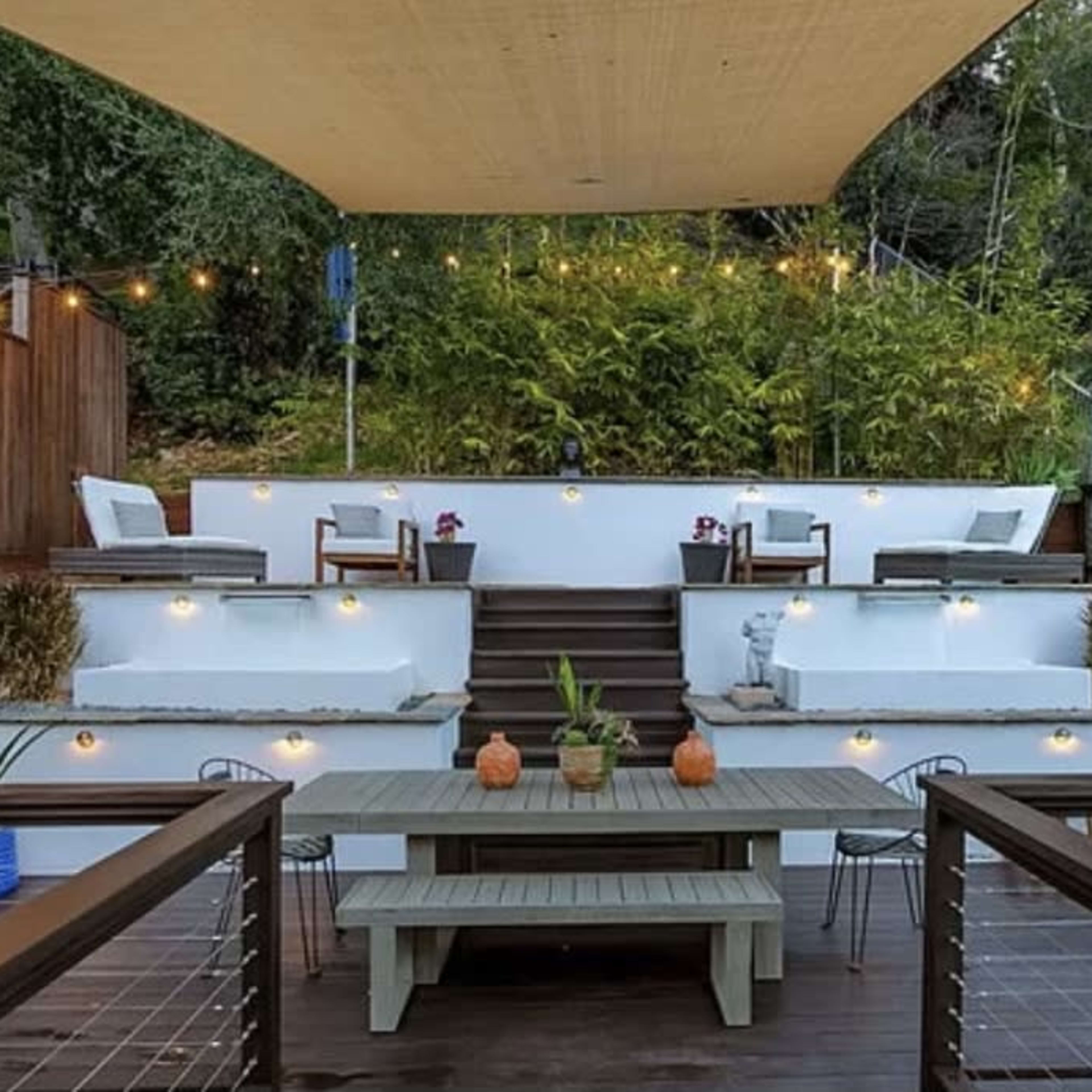 A modern outdoor patio area with a wooden dining table, decorative potted plants, and tiered seating areas surrounded by greenery and string lights.