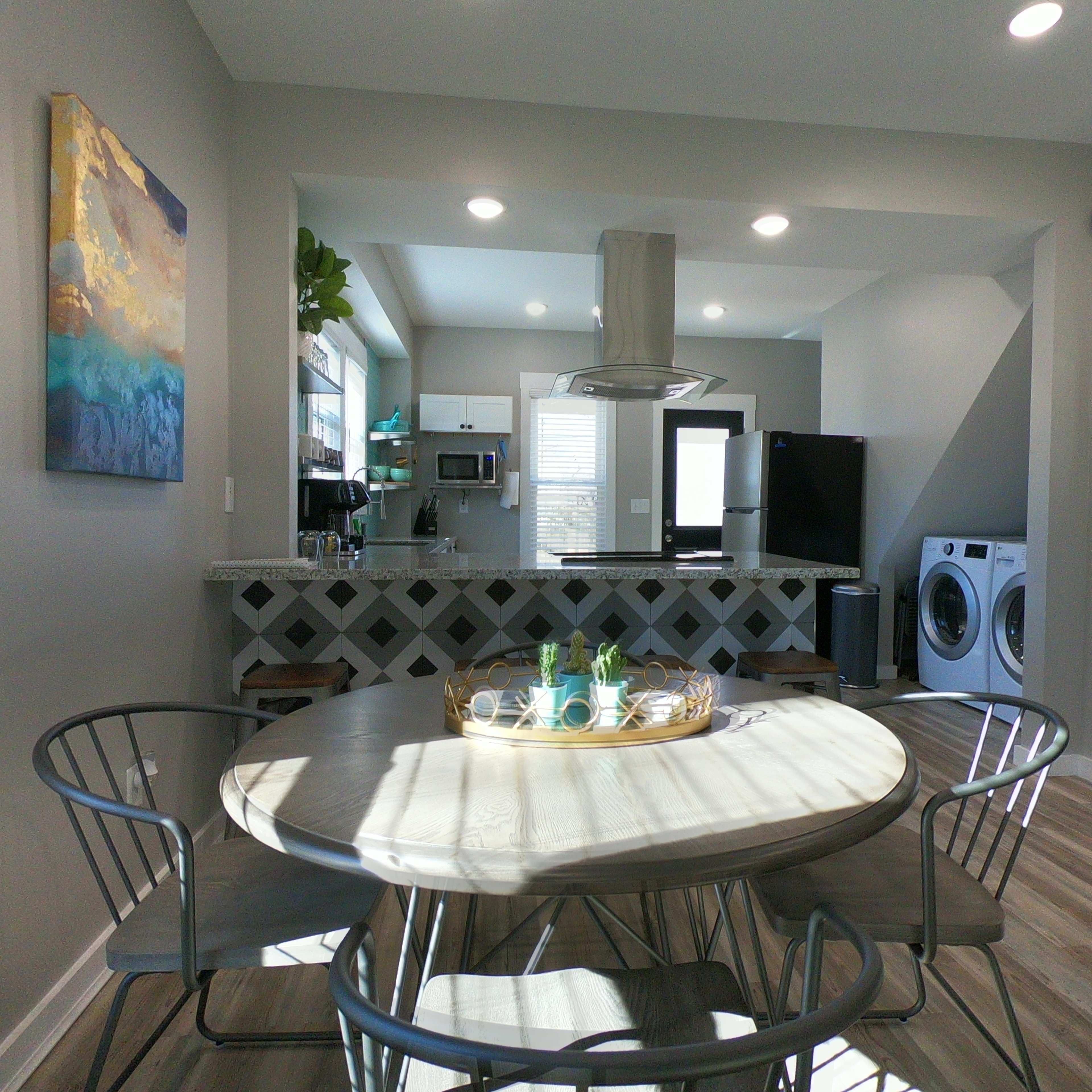 A modern kitchen and dining area features a round table with four chairs, a sleek countertop, and appliances in a well-lit space.