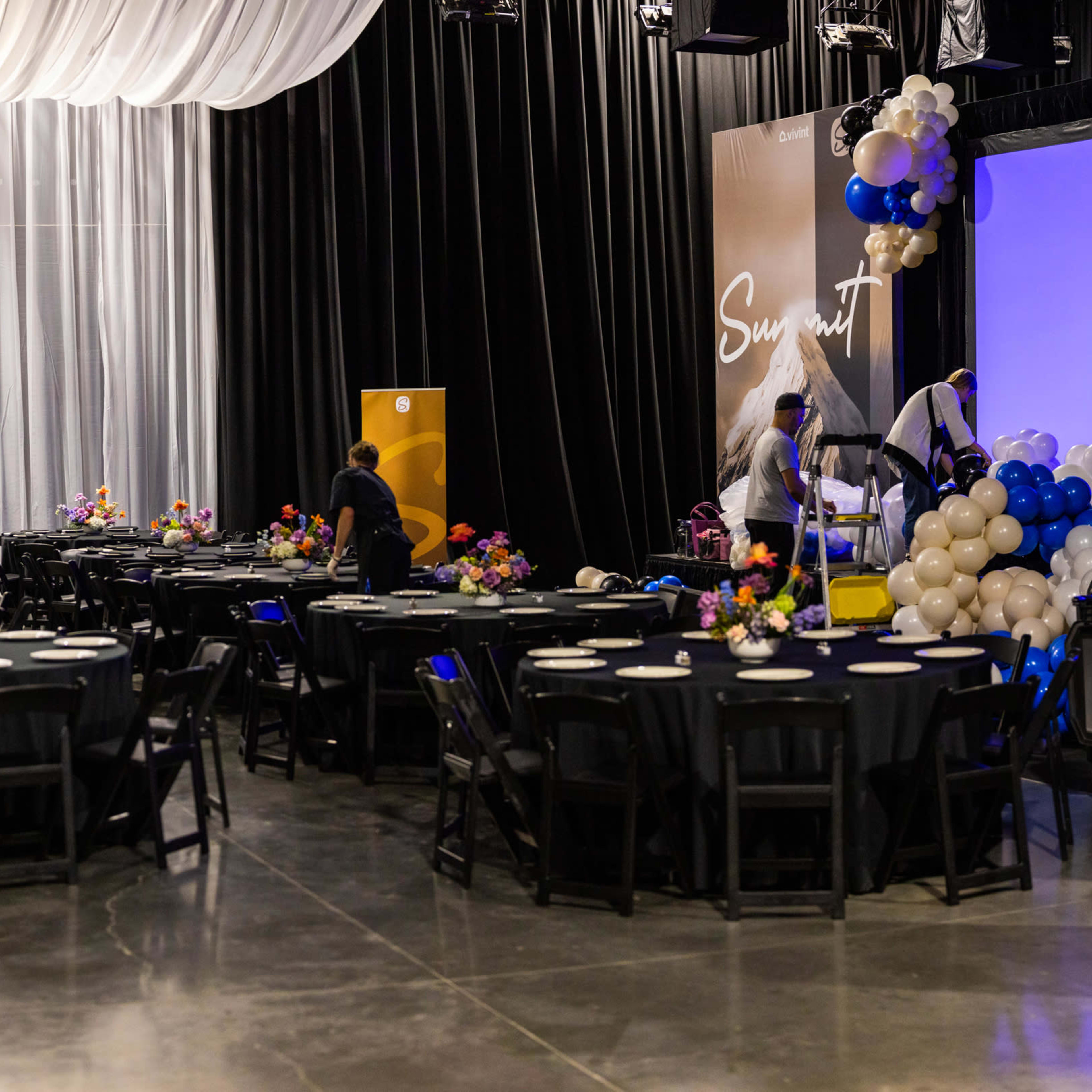 A spacious event hall is being prepared for a gathering, featuring several round tables adorned with floral centerpieces and a stage set up in the background.