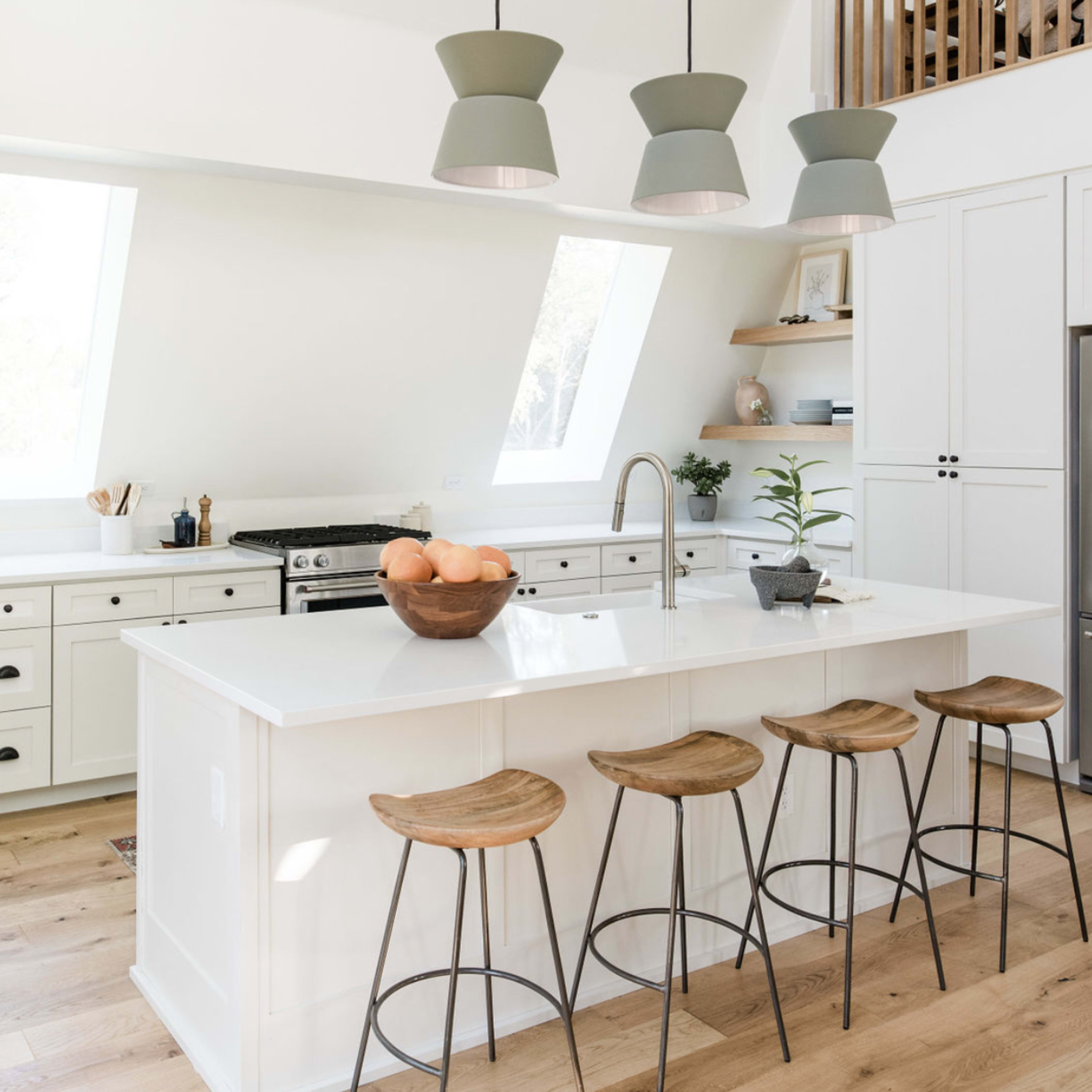 A modern kitchen with a white island, wooden stools, and large windows allowing natural light.