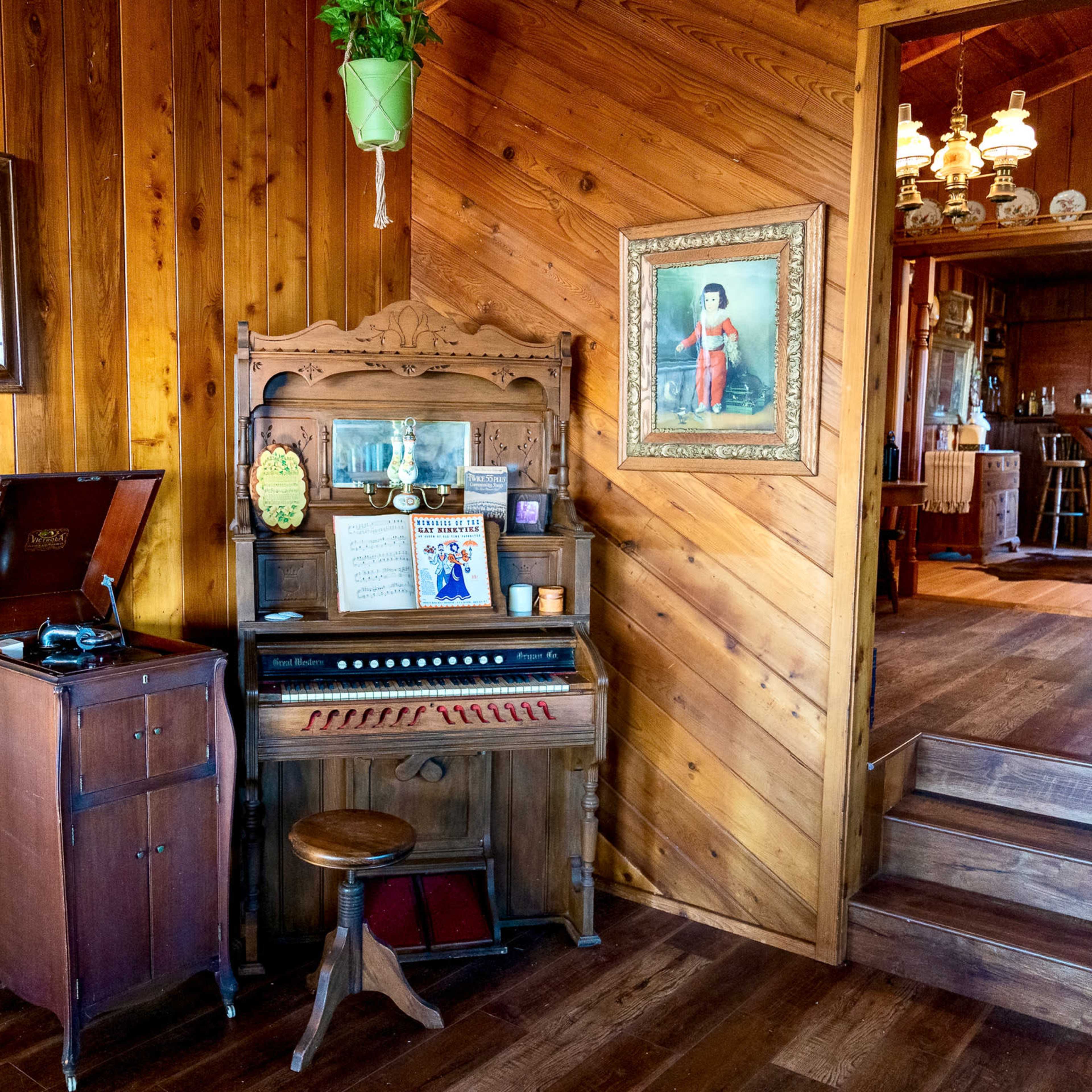 A rustic interior with wooden walls featuring an antique phonograph and a vintage organ, accompanied by framed artwork and a staircase leading to another room.