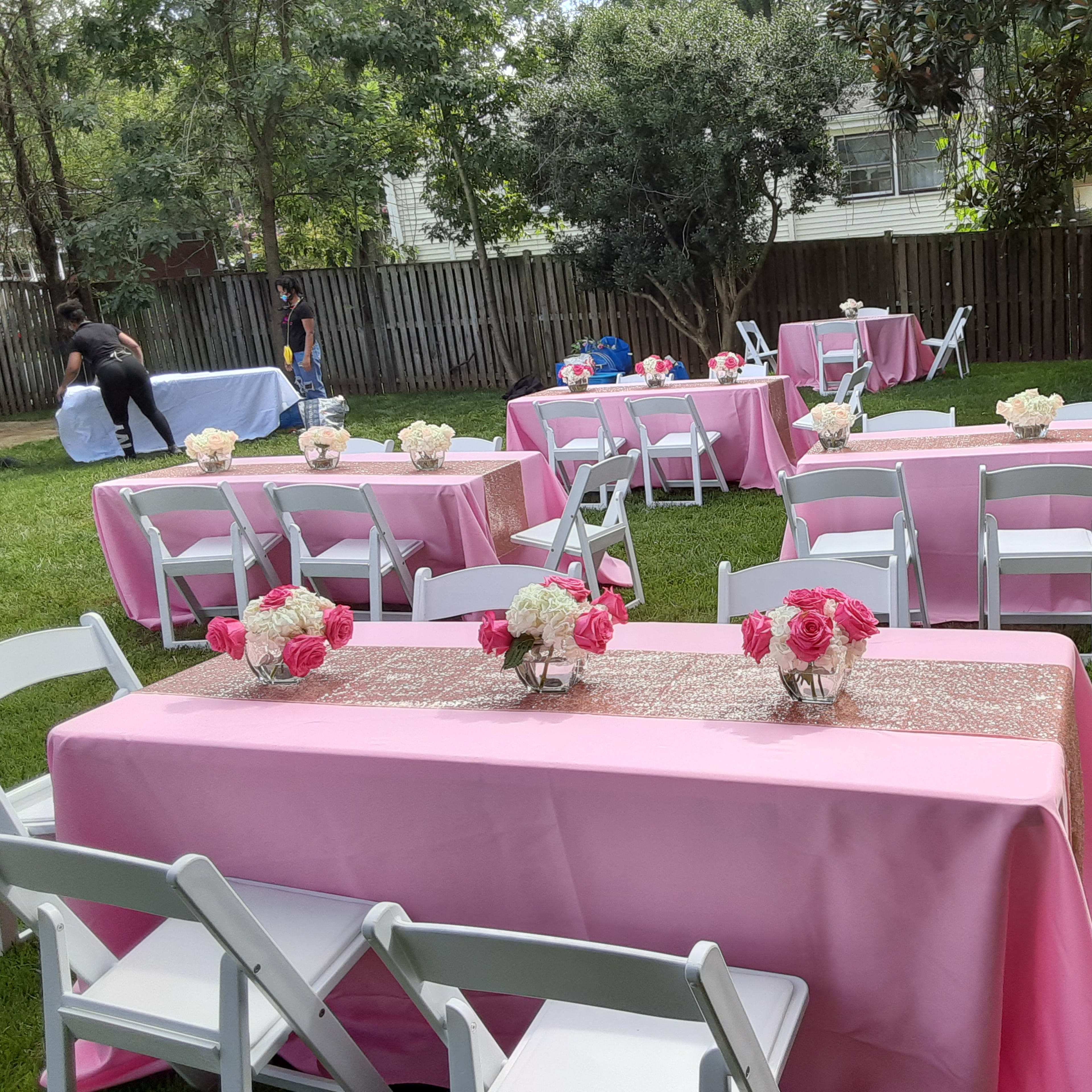 A garden setup with several tables covered in pink tablecloths and adorned with flower arrangements, surrounded by greenery.
