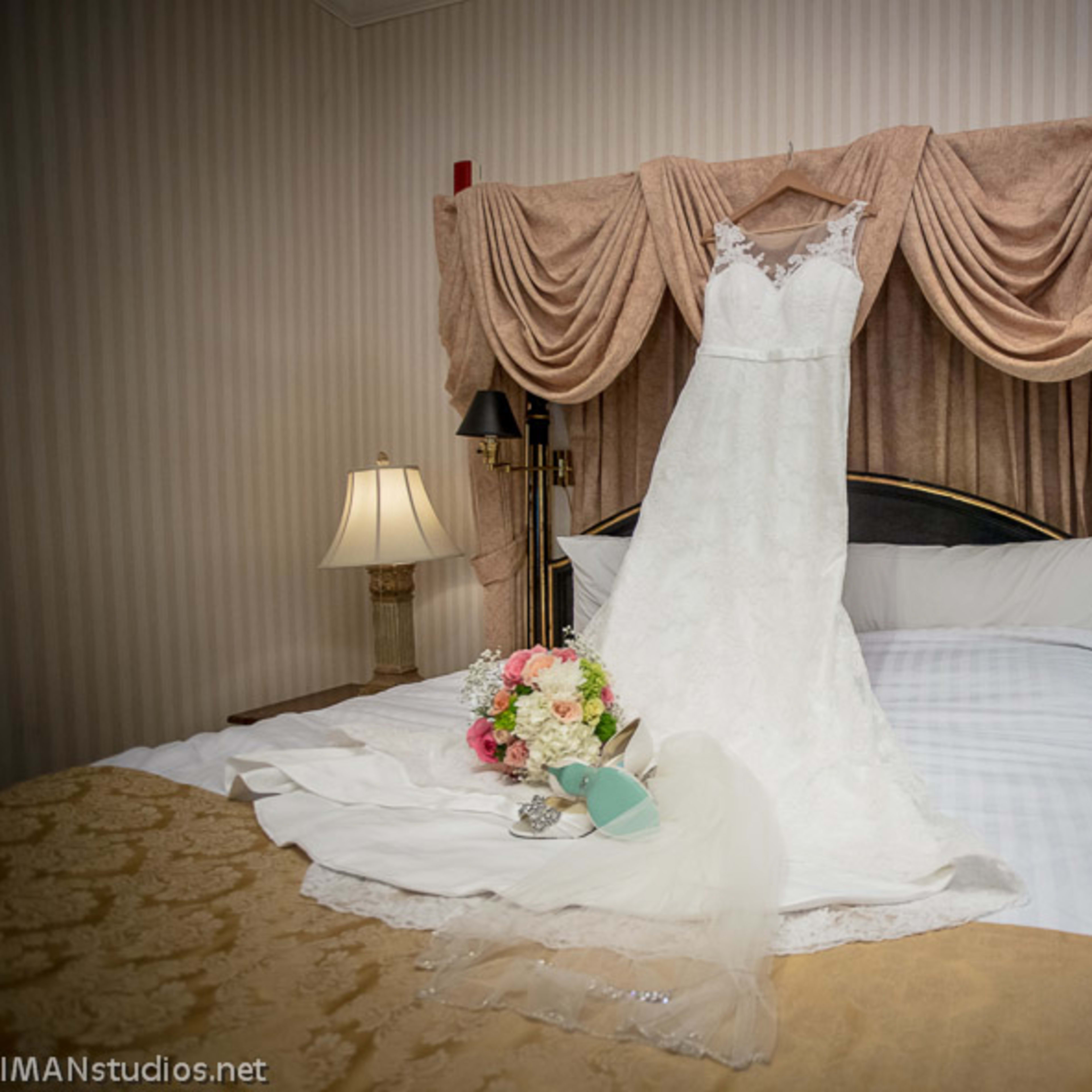 A wedding dress is displayed on a bed with a bouquet and accessories arranged nearby.