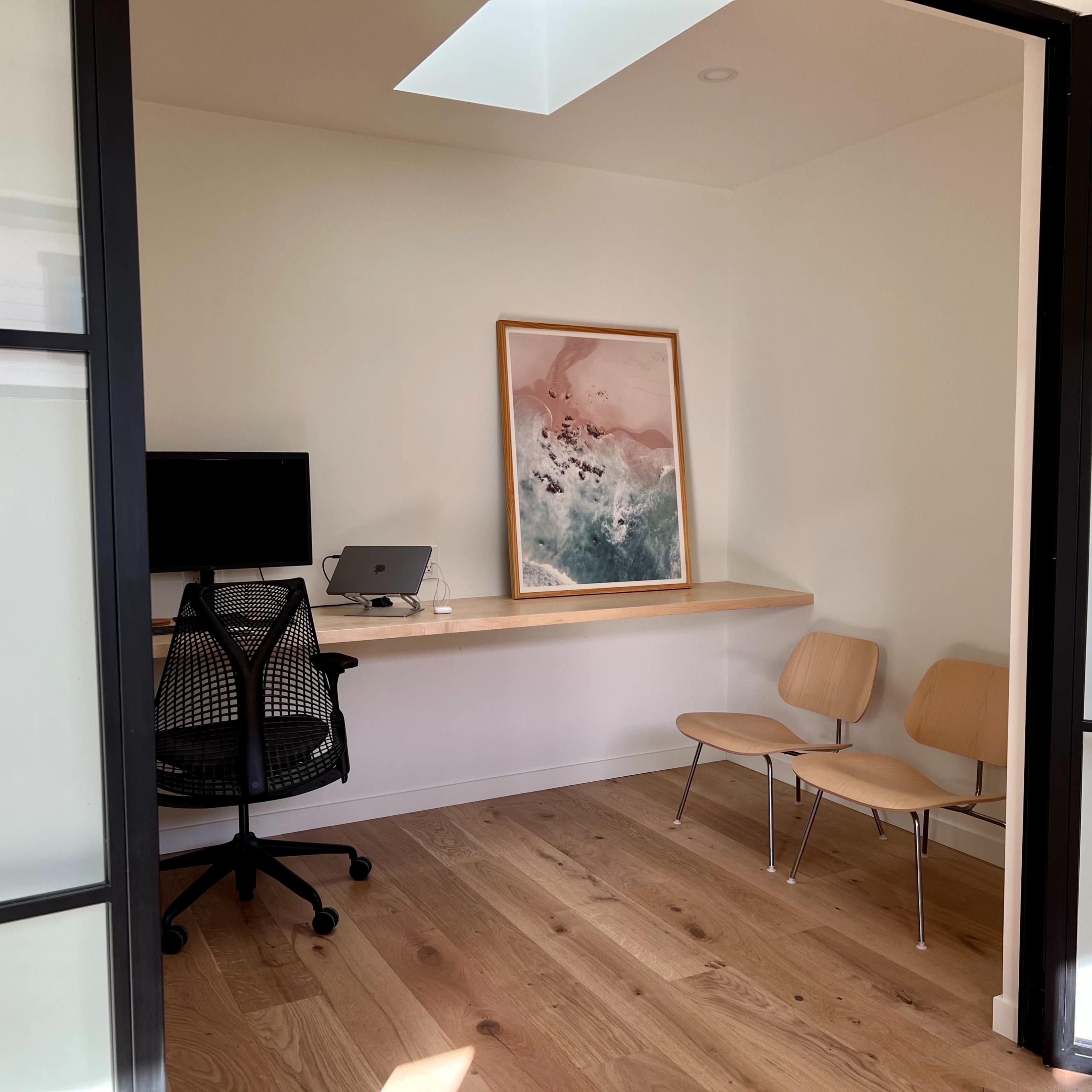 A minimalist office space with a desk, a computer monitor, and two wooden chairs.
