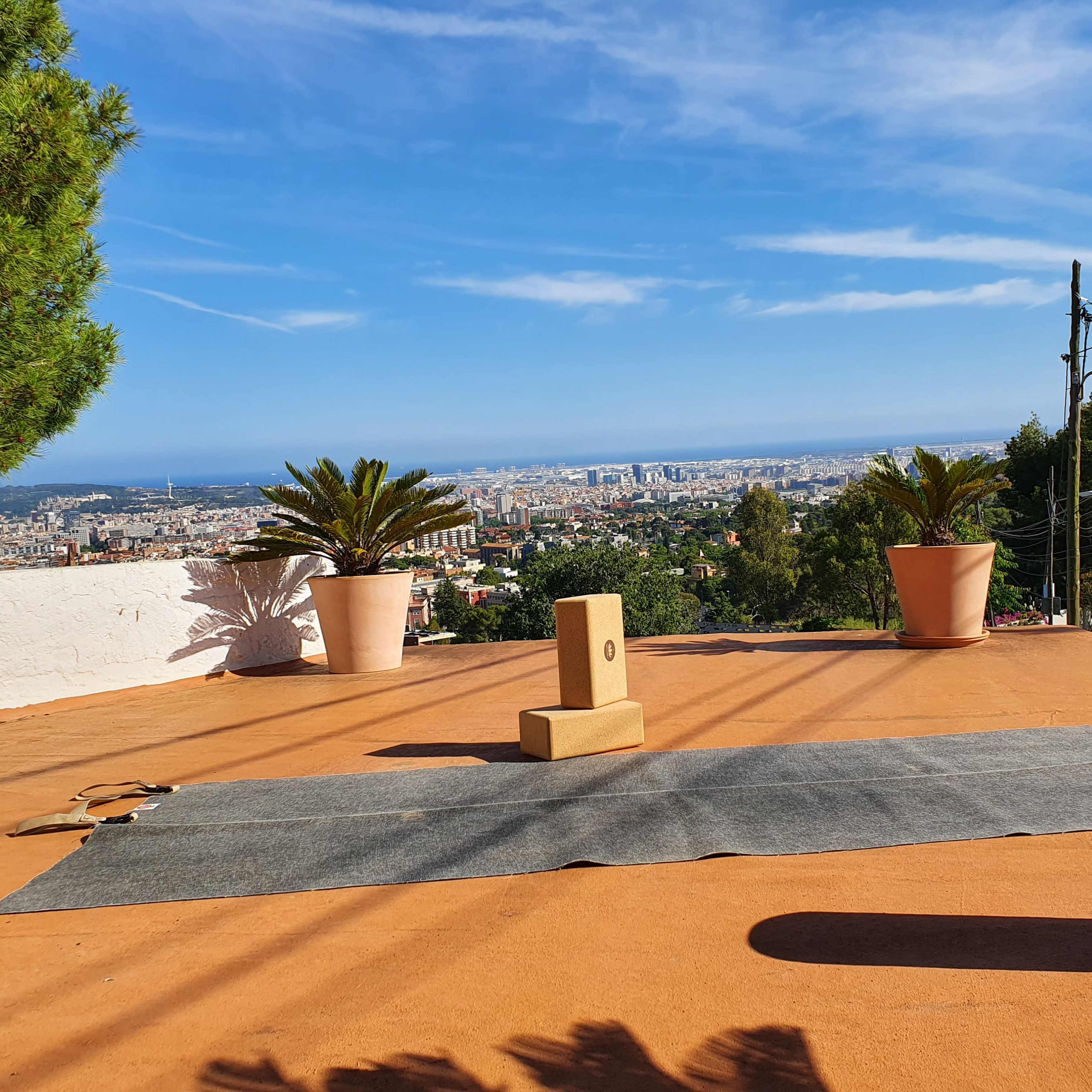 A yoga mat and props are set up on a terrace overlooking a cityscape with palm trees and clear blue skies.