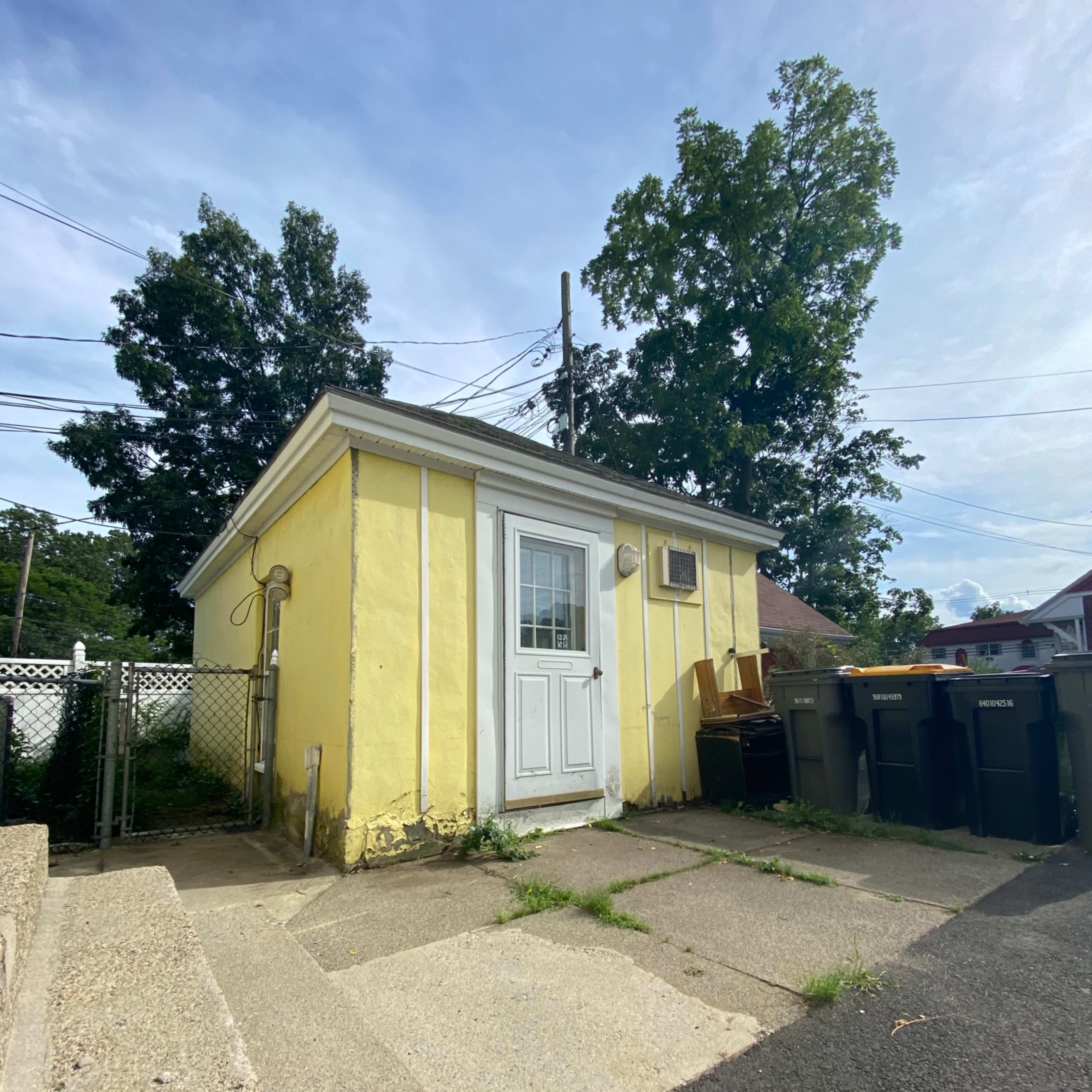 A small yellow shed with a white door and an air conditioning unit sits next to a pathway and several trash bins.