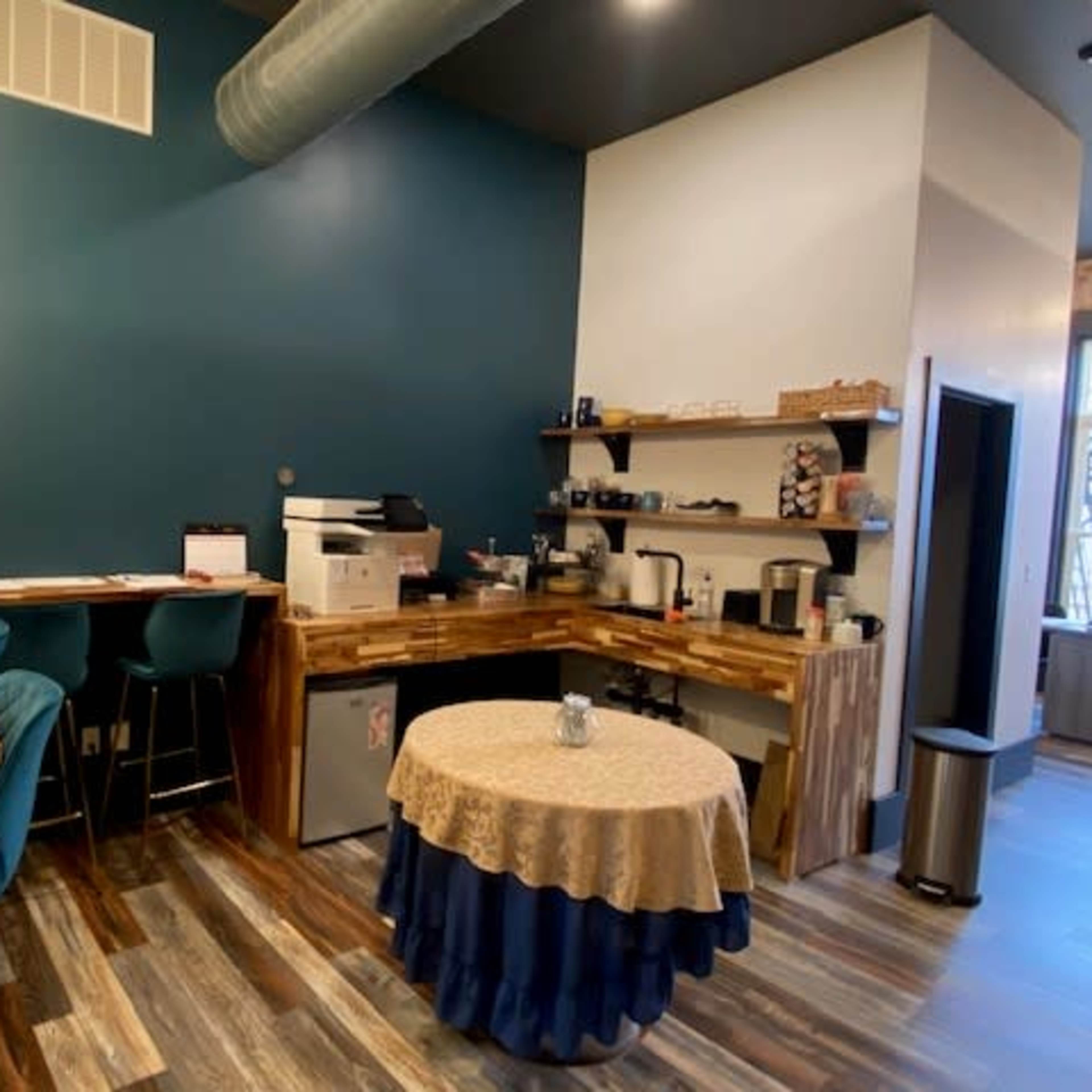 A small coffee station with a table draped in a lace tablecloth is set against a teal wall, featuring wooden countertops and various kitchen appliances.
