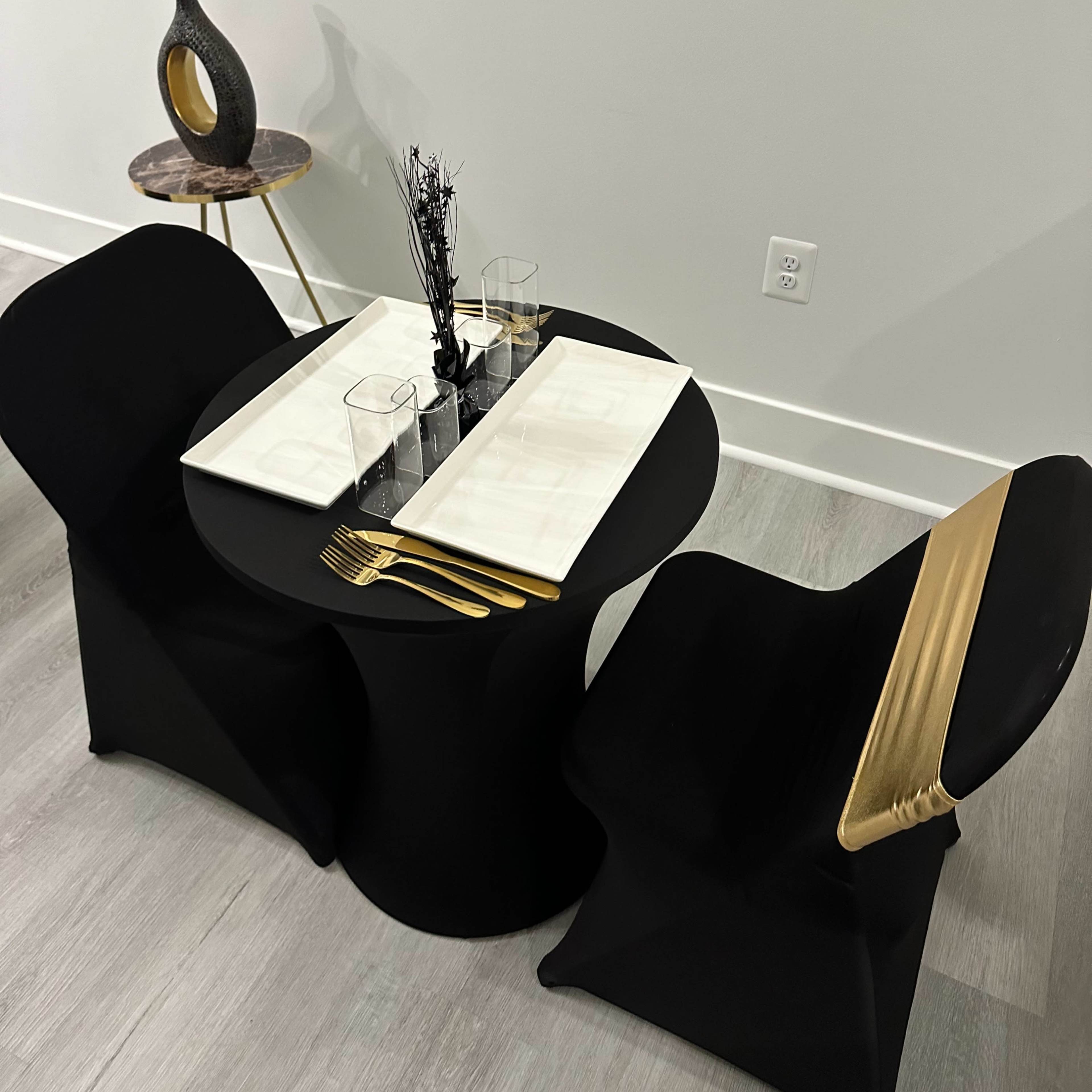 A modern dining setup features a black table and two chairs with a gold accent, complemented by a minimalist centerpiece and glassware.