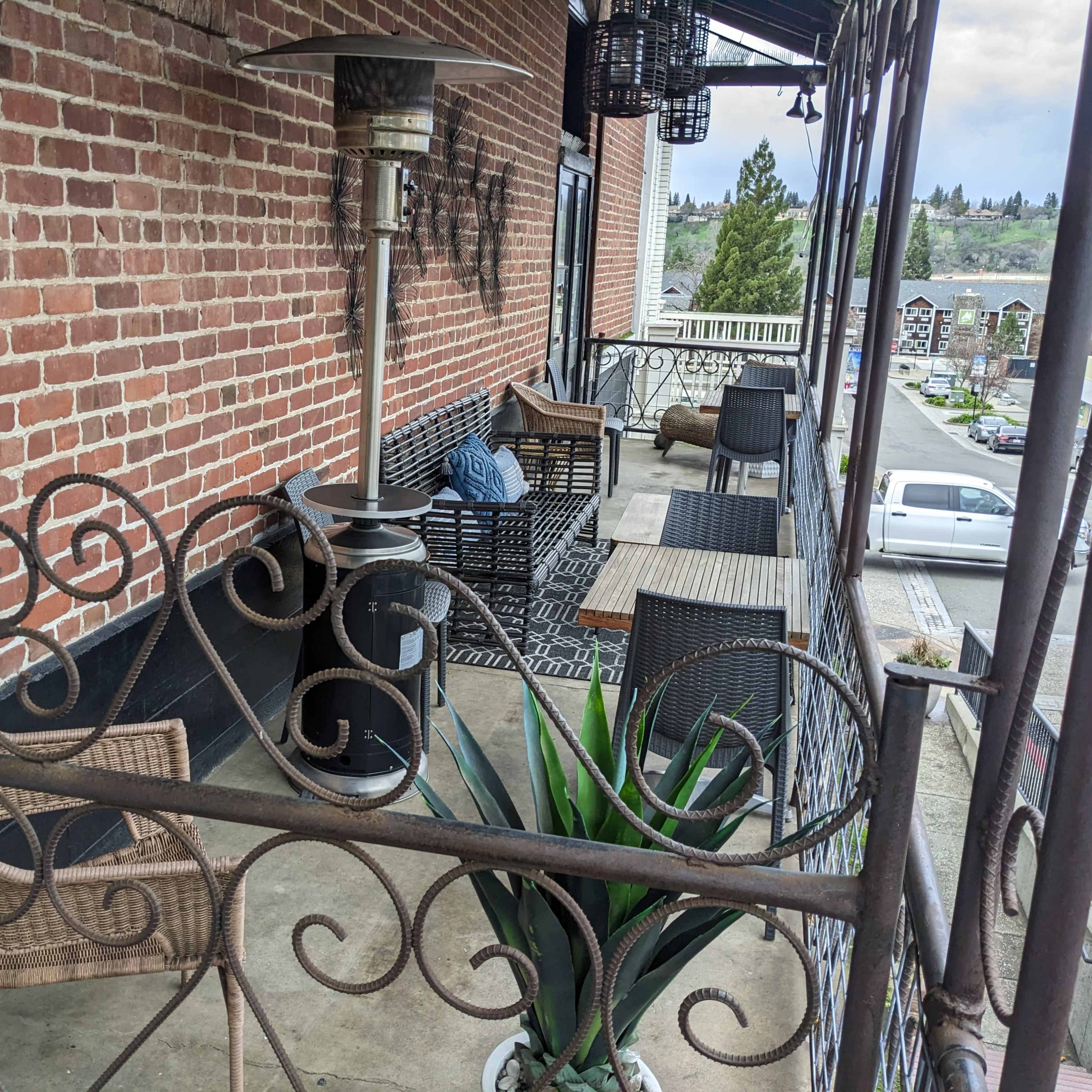 A covered patio with outdoor furniture, including a metal table, woven chairs, and a heater, adjacent to a brick wall.