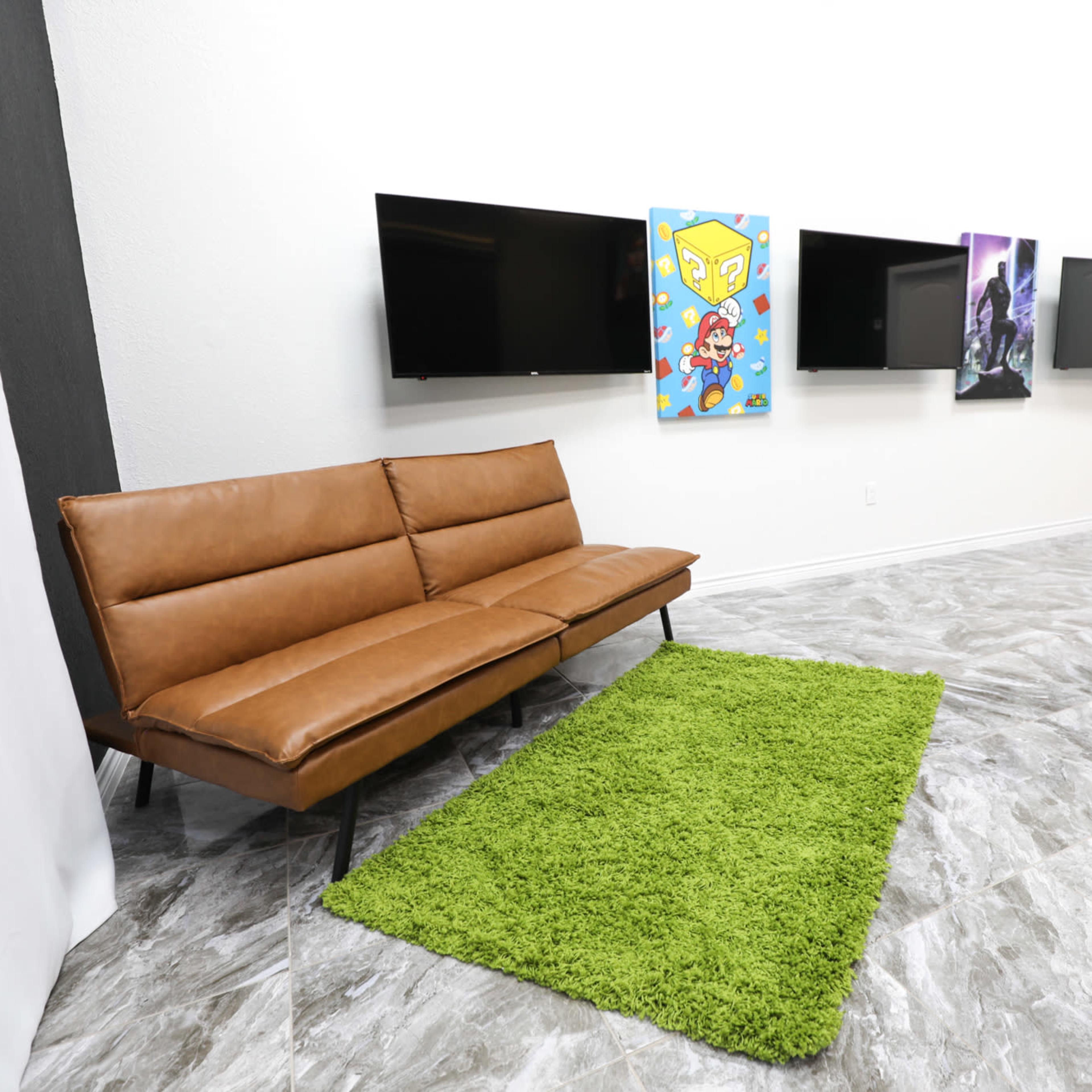 A modern interior space with a brown couch, a green rug, and multiple framed artworks displayed on the wall opposite two flat-screen televisions.