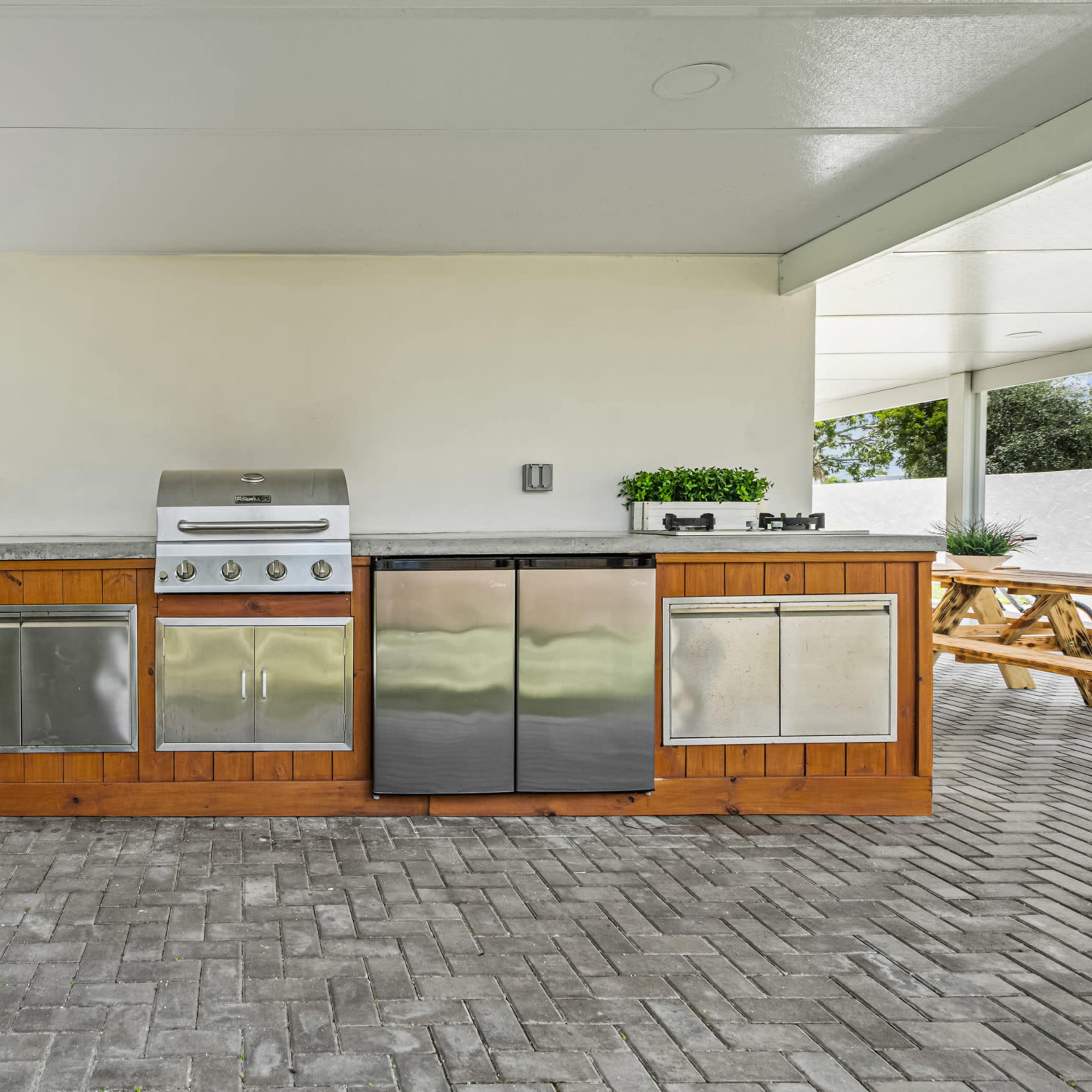 A modern outdoor kitchen featuring a stainless steel grill, a refrigerator, and storage cabinets, all under a covered patio.