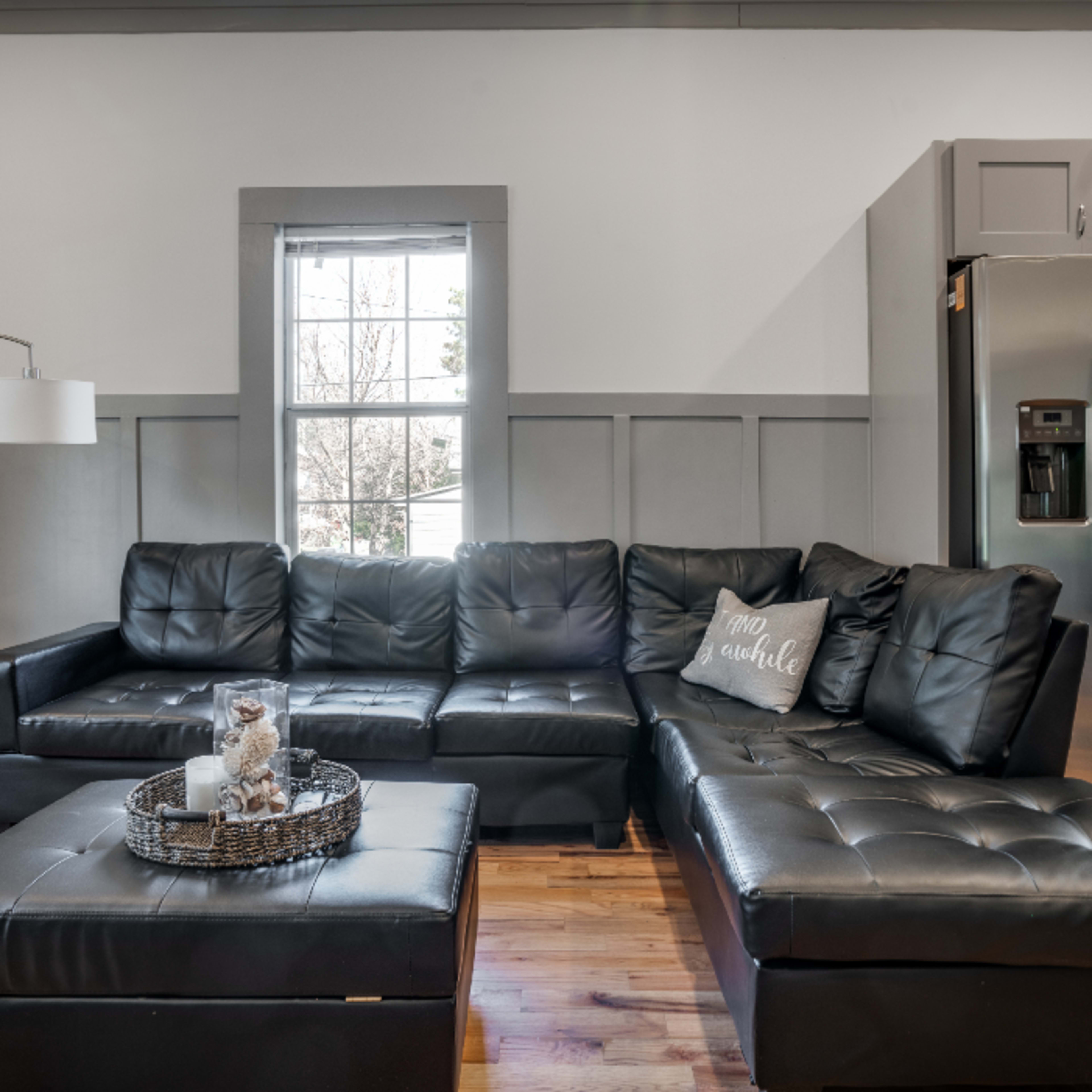 A modern living room featuring a black leather sectional sofa, a coffee table, and a stainless steel refrigerator in the corner.