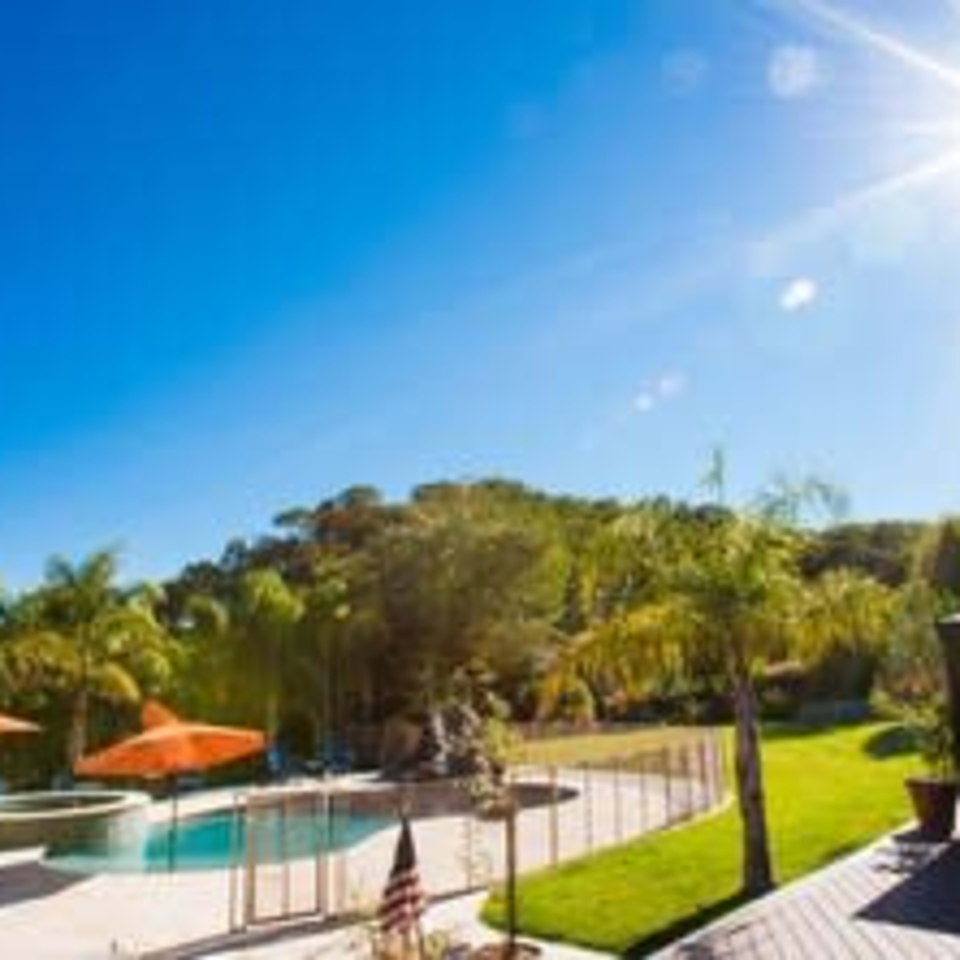 A bright, sunny backyard features a swimming pool, palm trees, umbrellas, and a gazebo.