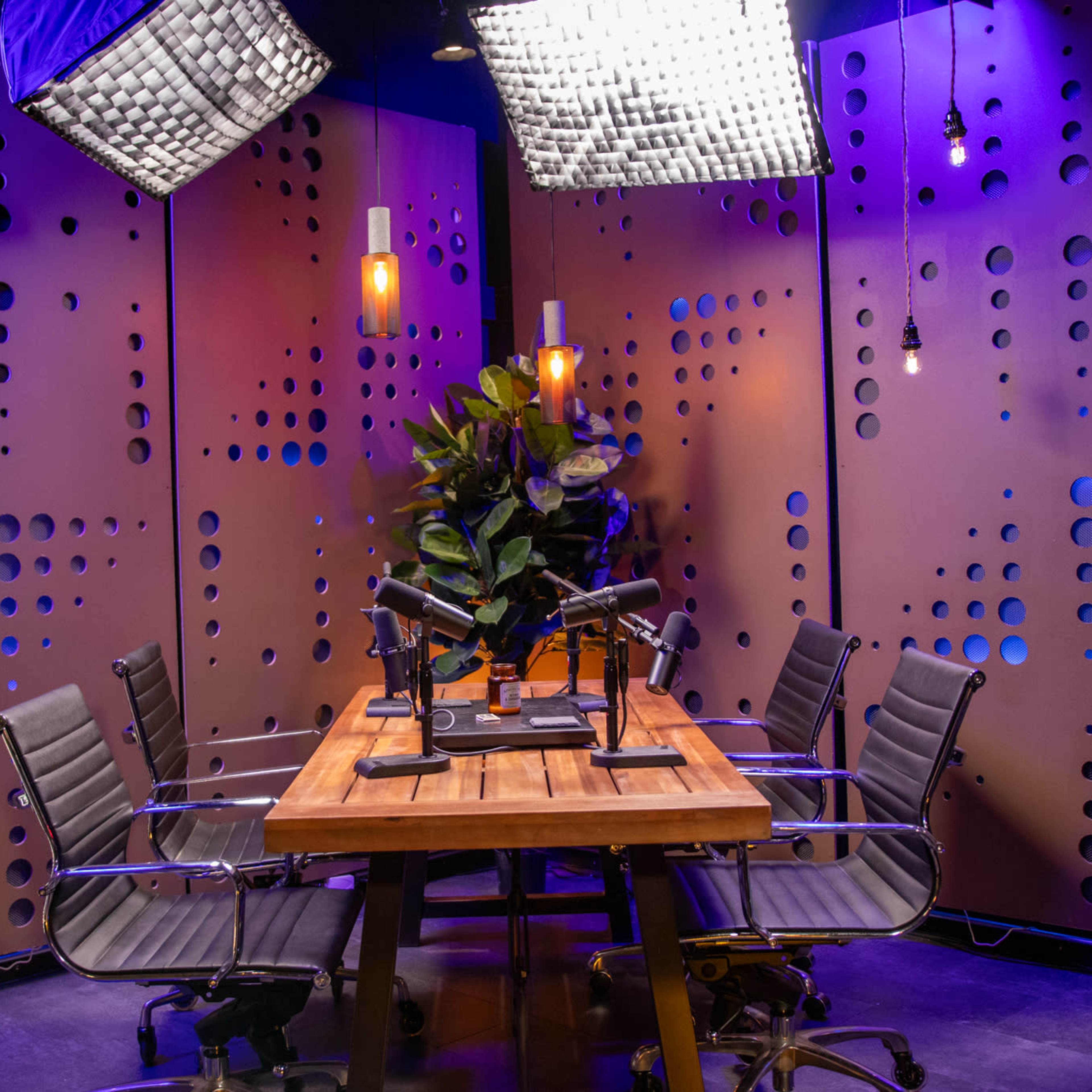 A modern podcast studio features a wooden table surrounded by sleek chairs, with microphones set up and overhead lights illuminating the space.